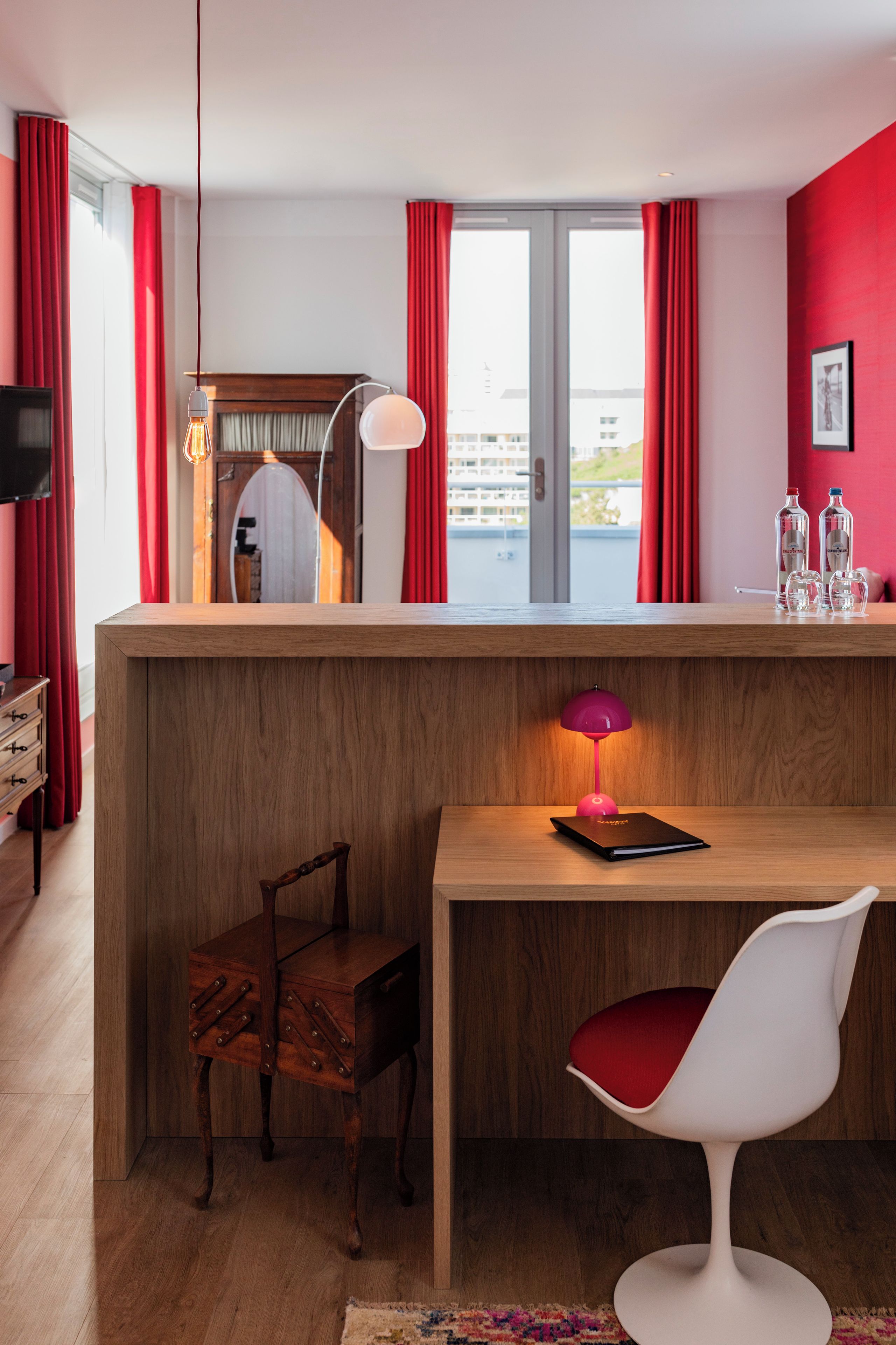 A modern room with a wooden desk, a red chair, a small table, a lamp, and a view through a window with red curtains.
