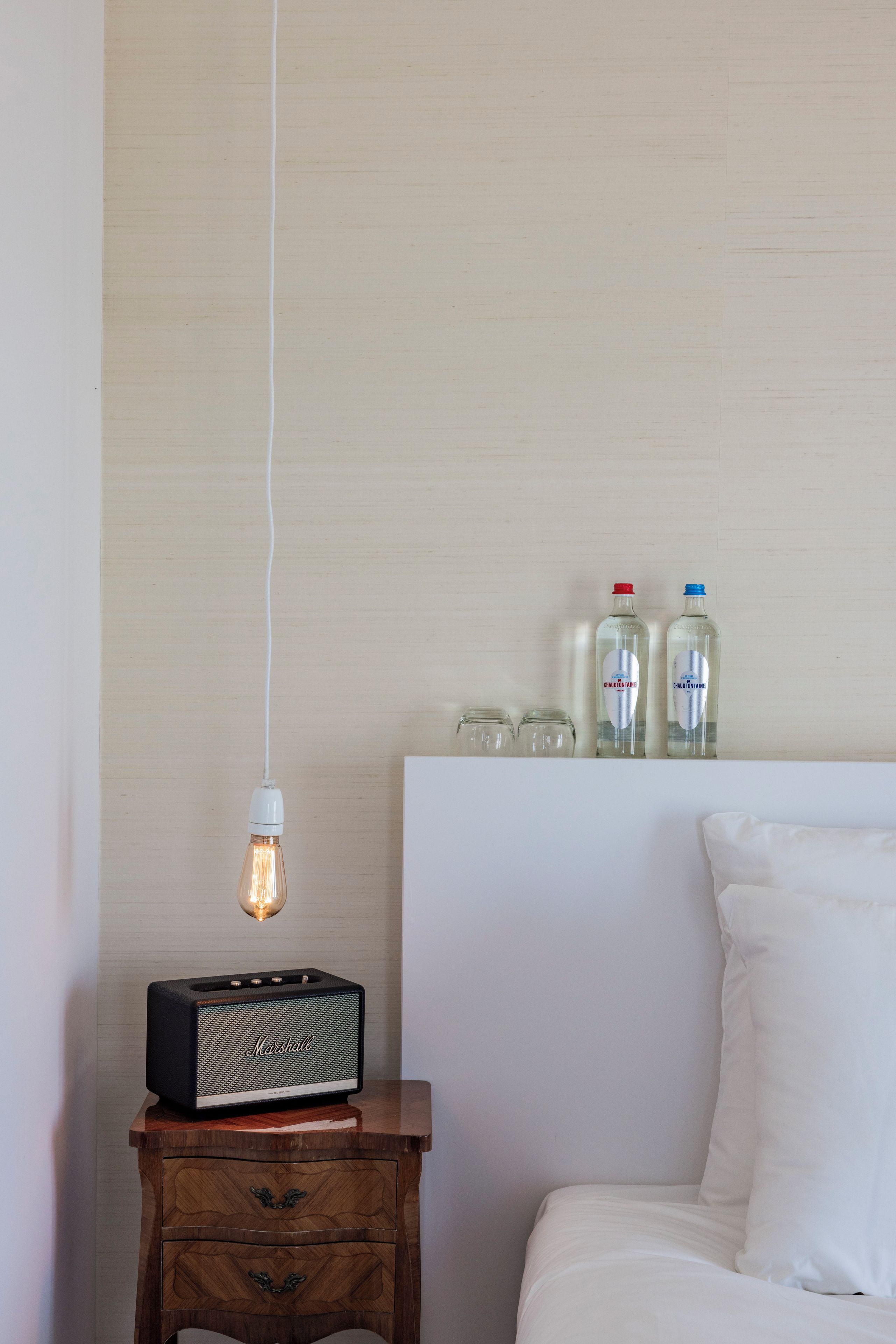Een slaapkamer met een houten nachtkastje, een vintage radio, een hangende lamp en twee flessen op het hoofdeinde.