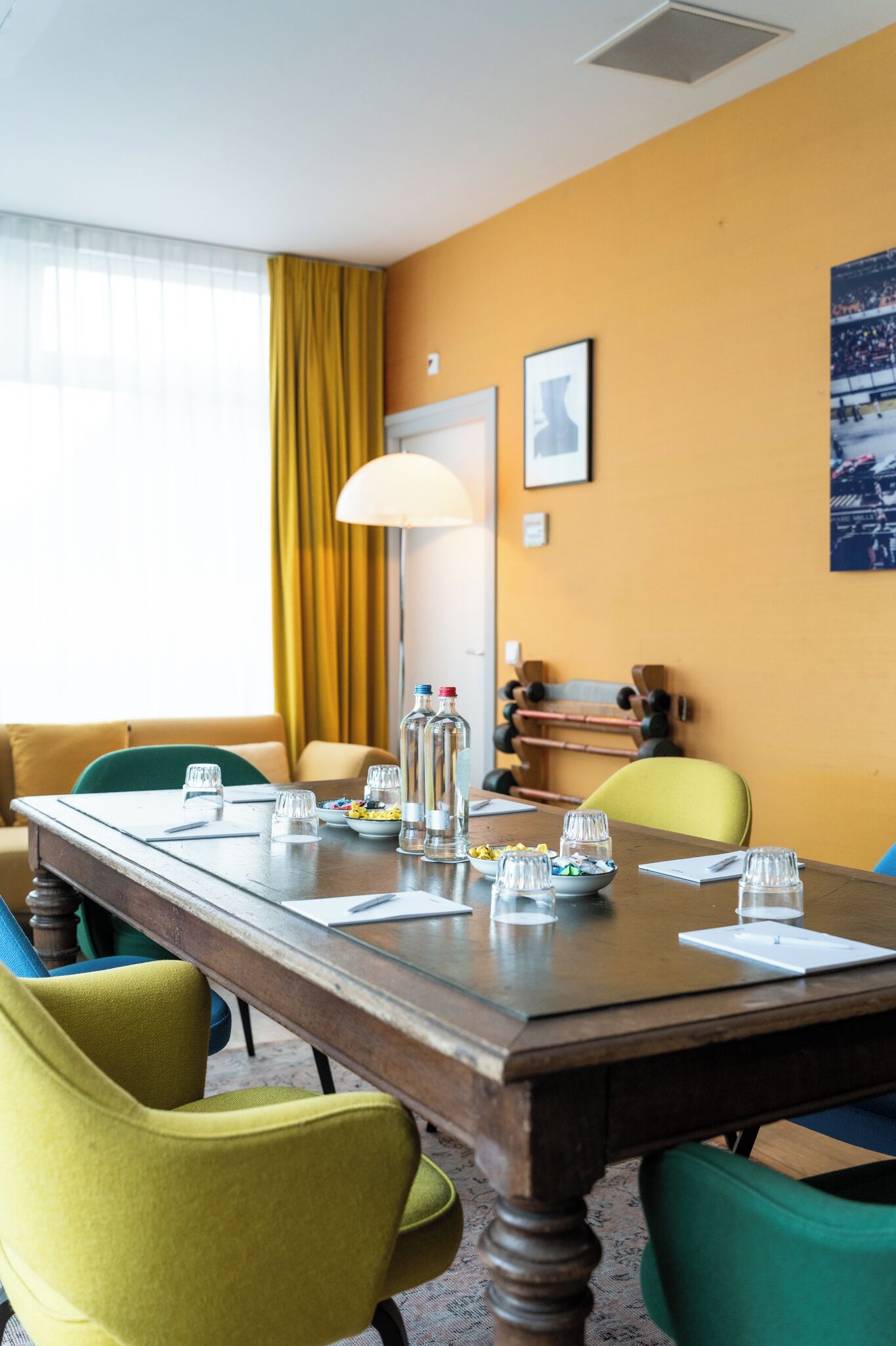 A hotel room with a wooden dining table set for four, yellow chairs, a lamp, and a window with yellow curtains.
