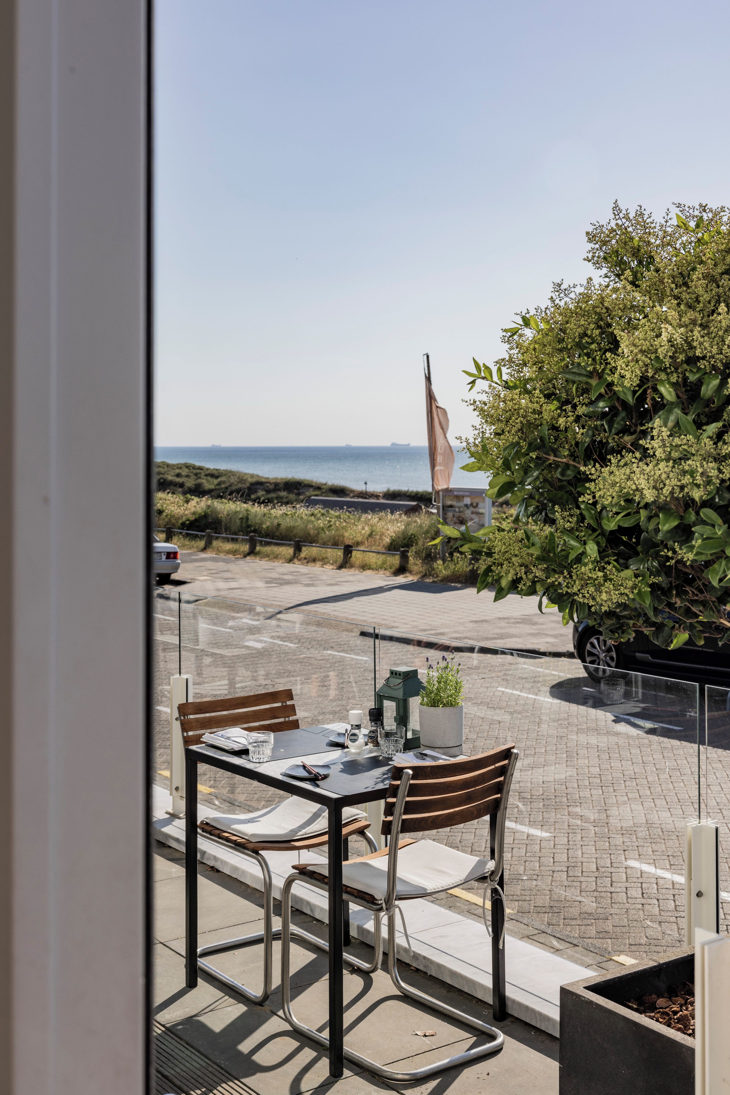 A table with chairs and a potted plant, overlooking a paved area with a flag and a view of the sea.