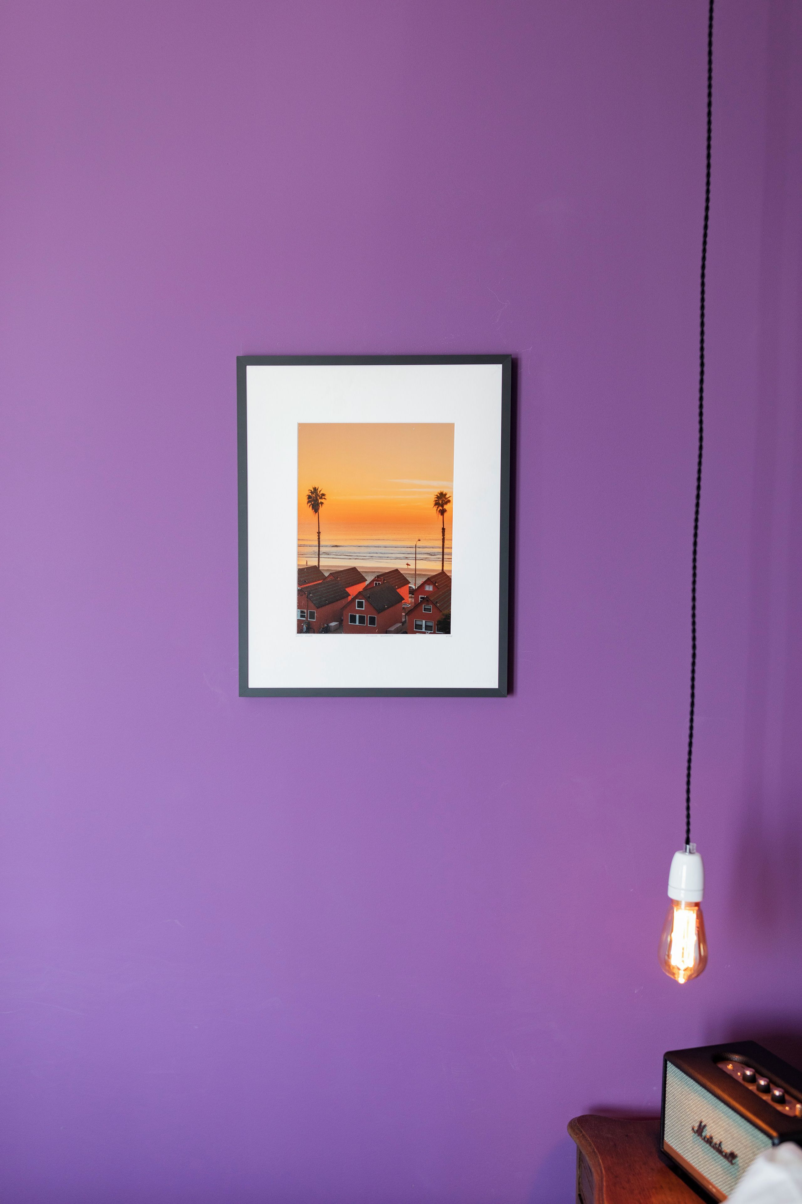 Une photographie encadrée représentant un coucher de soleil sur une plage bordée de palmiers et de maisons, accrochée à un mur violet.