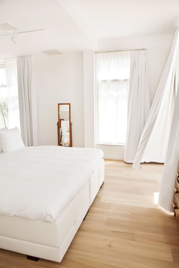 A modern bedroom with a large bed, a nightstand, a mirror, and sheer curtains. - Vesper Hotel (Noordwijk)