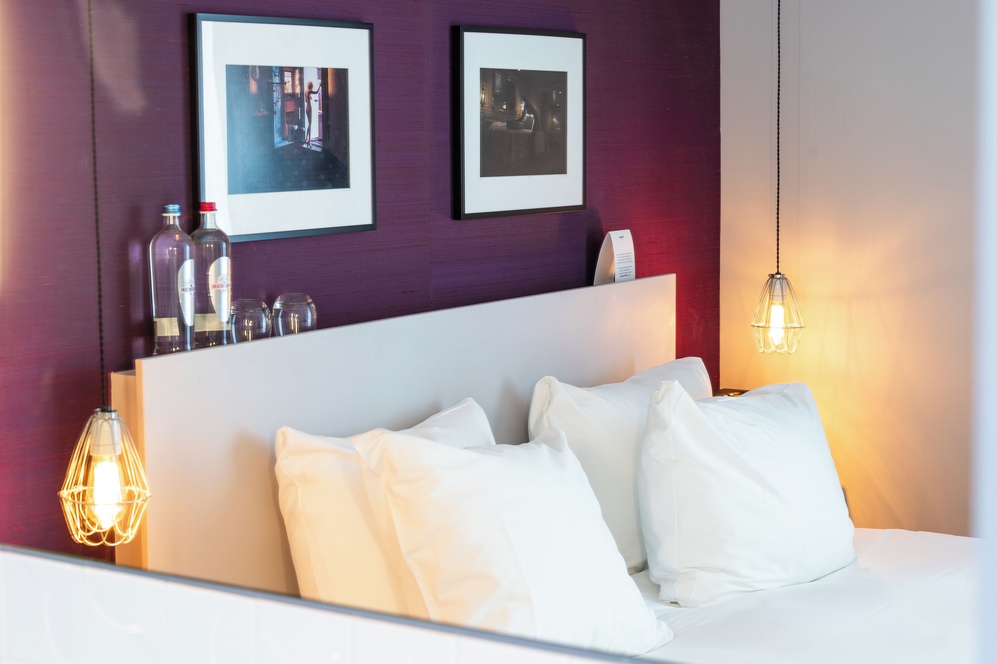 Habitación de hotel con cama, almohadas blancas, cabecero, dos cuadros enmarcados en la pared y una lámpara colgante.