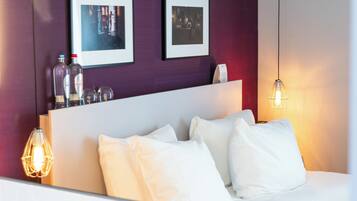 Habitación de hotel con cama, almohadas blancas, cabecero, dos cuadros enmarcados en la pared y una lámpara colgante.