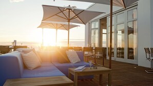 A sunlit outdoor seating area with a sofa, tables, and umbrellas, overlooking a body of water. - Vesper Hotel (Noordwijk)