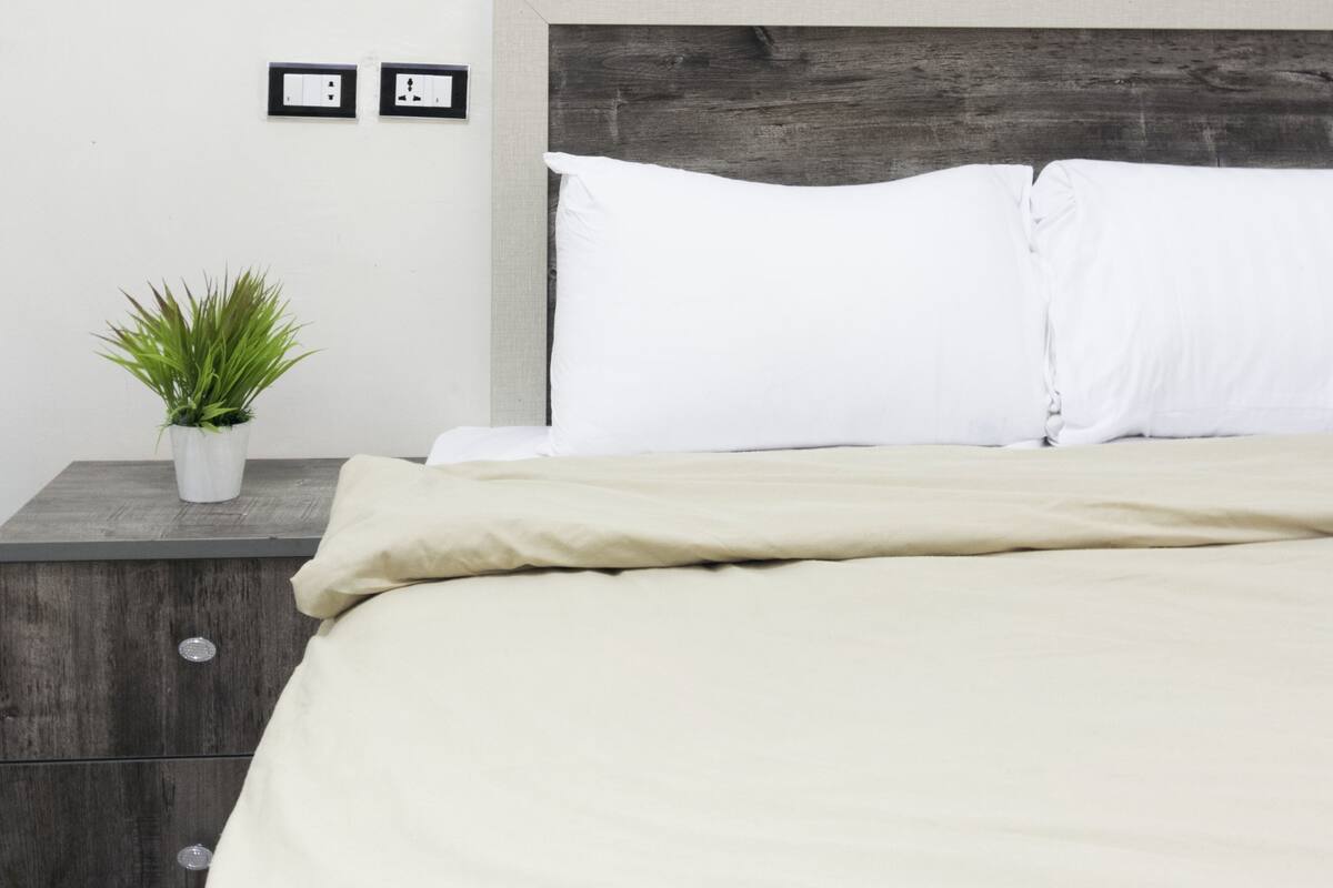 Ein Bett mit hölzernem Kopfteil, weißen Kissen und einer beigen Decke. Auf einem Nachttisch neben dem Bett steht eine kleine Topfpflanze.