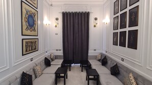 A room with a high ceiling, a ceiling fan, a large window with dark curtains, a sofa with matching cushions, and a wall adorned with framed pictures. - The Australian Hostel (Cairo)
