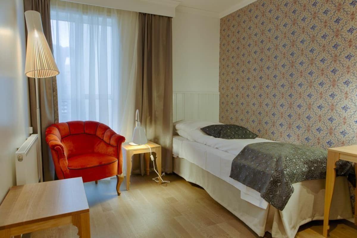 Habitación de hotel pequeña con una cama individual, un sillón rojo, una mesita de noche con lámpara y un papel tapiz estampado.
