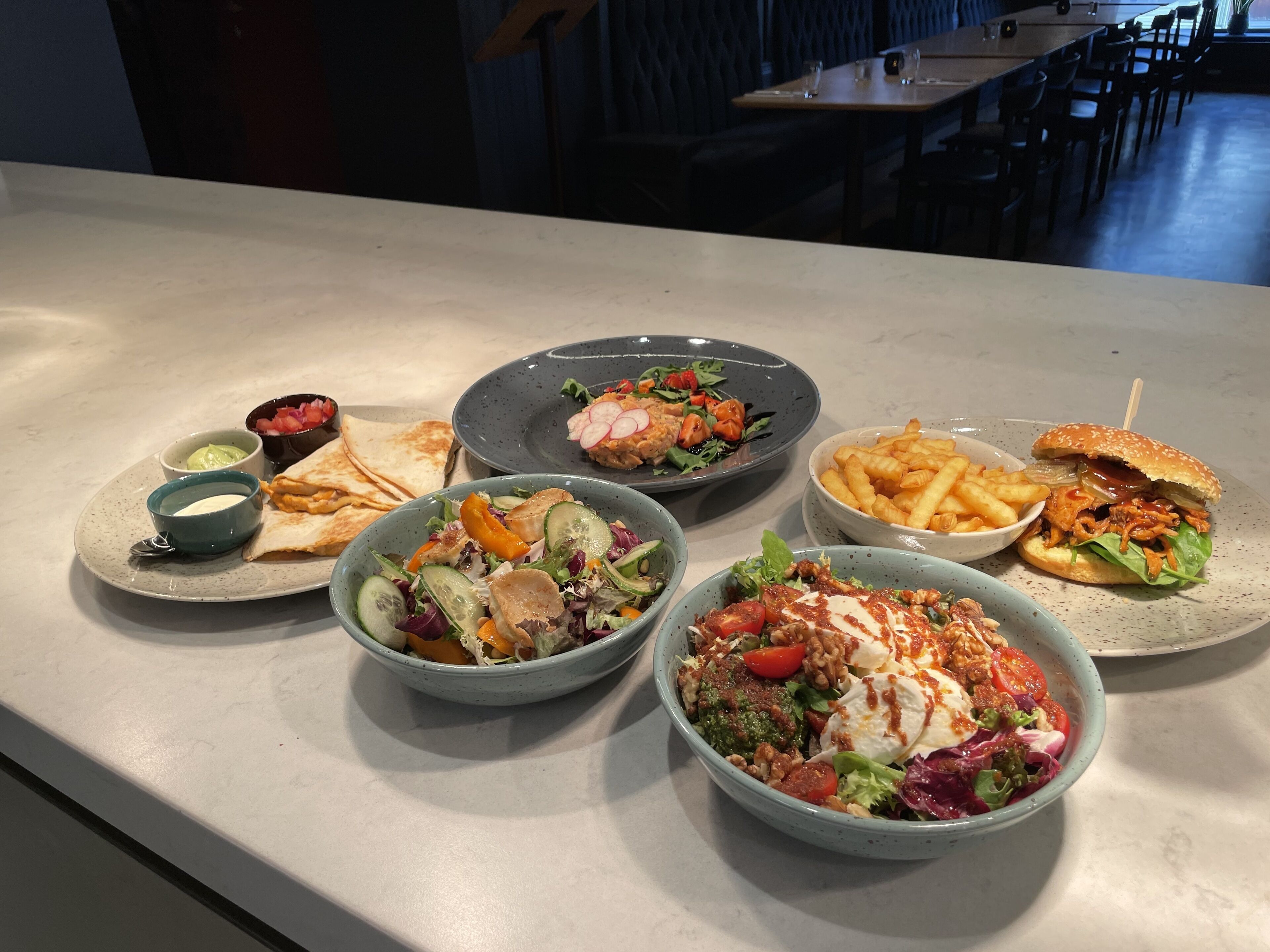Una mesa de comedor con diversos platos, entre ellos ensaladas, sándwiches y papas fritas.