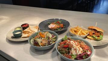 Una mesa de comedor con diversos platos, entre ellos ensaladas, sándwiches y papas fritas.