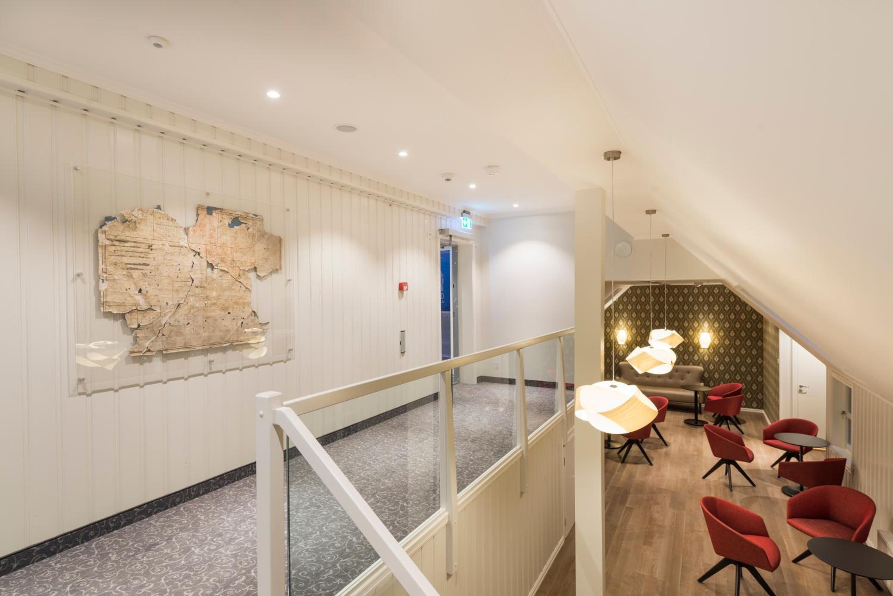 A hotel lobby with a modern design, featuring a staircase, seating area with red chairs, and a decorative wall piece.