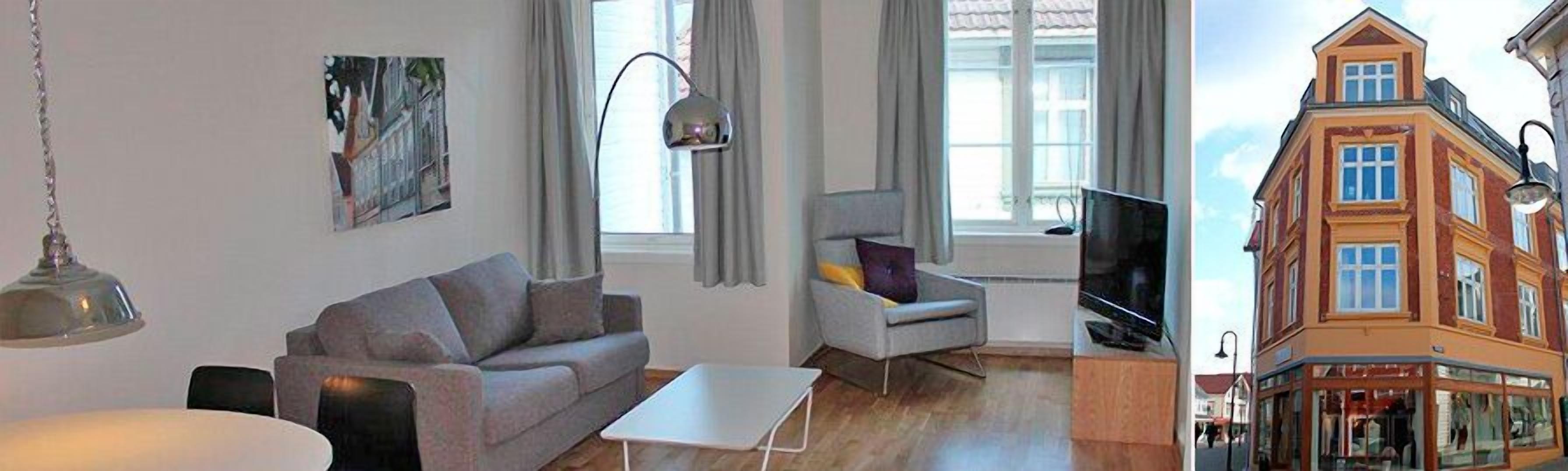 Ein modernes Wohnzimmer mit einer Couch, einem Sessel und einem Fernseher, großen Fenstern und Blick auf eine Straße mit Gebäuden.