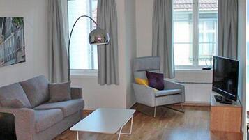 Ein modernes Wohnzimmer mit einer Couch, einem Sessel und einem Fernseher, großen Fenstern und Blick auf eine Straße mit Gebäuden.