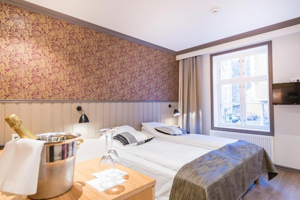 A hotel room with a bed, a bedside table with a bottle of champagne, a lamp, and a window with curtains.