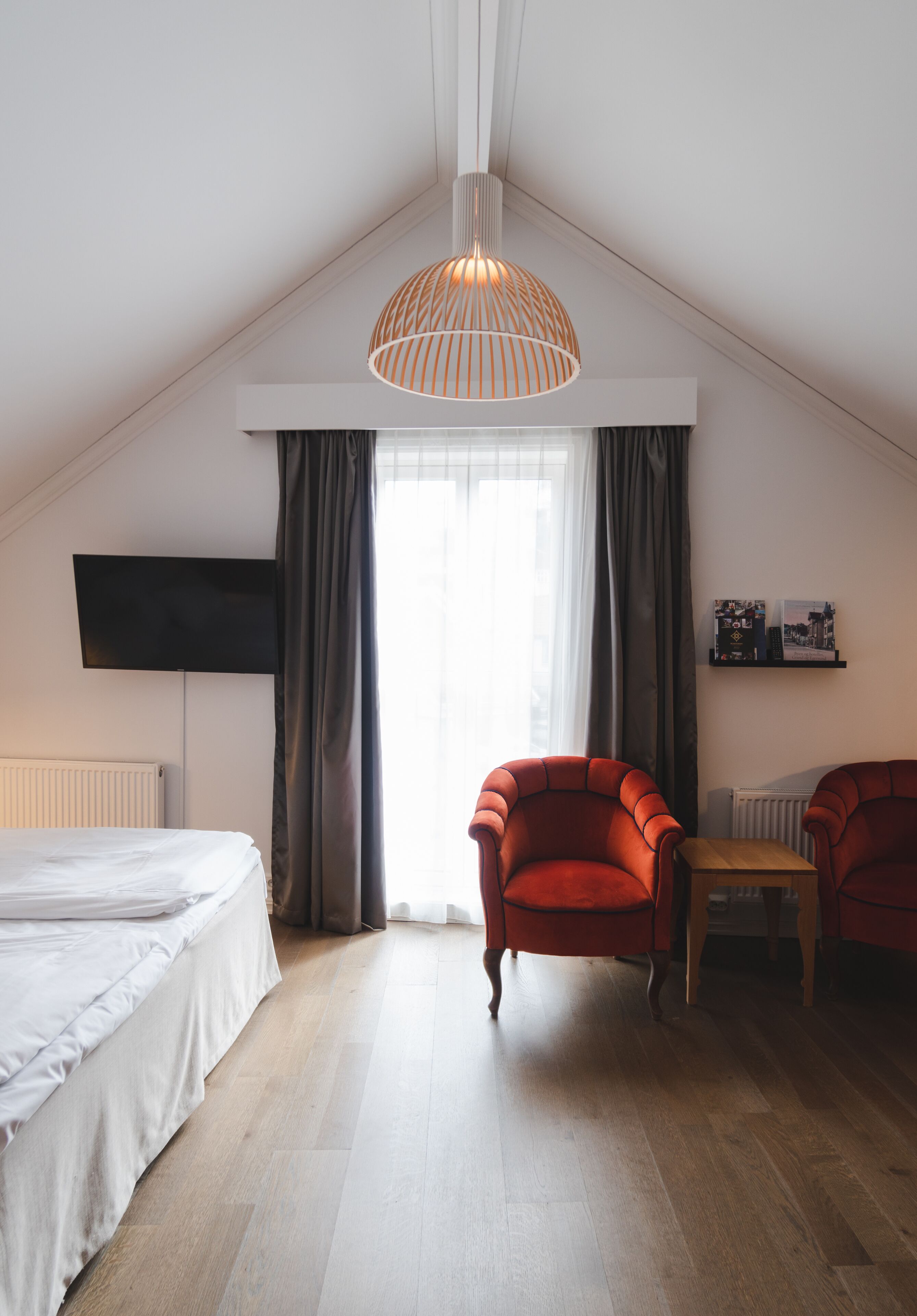 Un dormitorio con una cama, dos sillones rojos, una mesa de madera, un televisor montado en la pared y un amplio ventanal con cortinas.