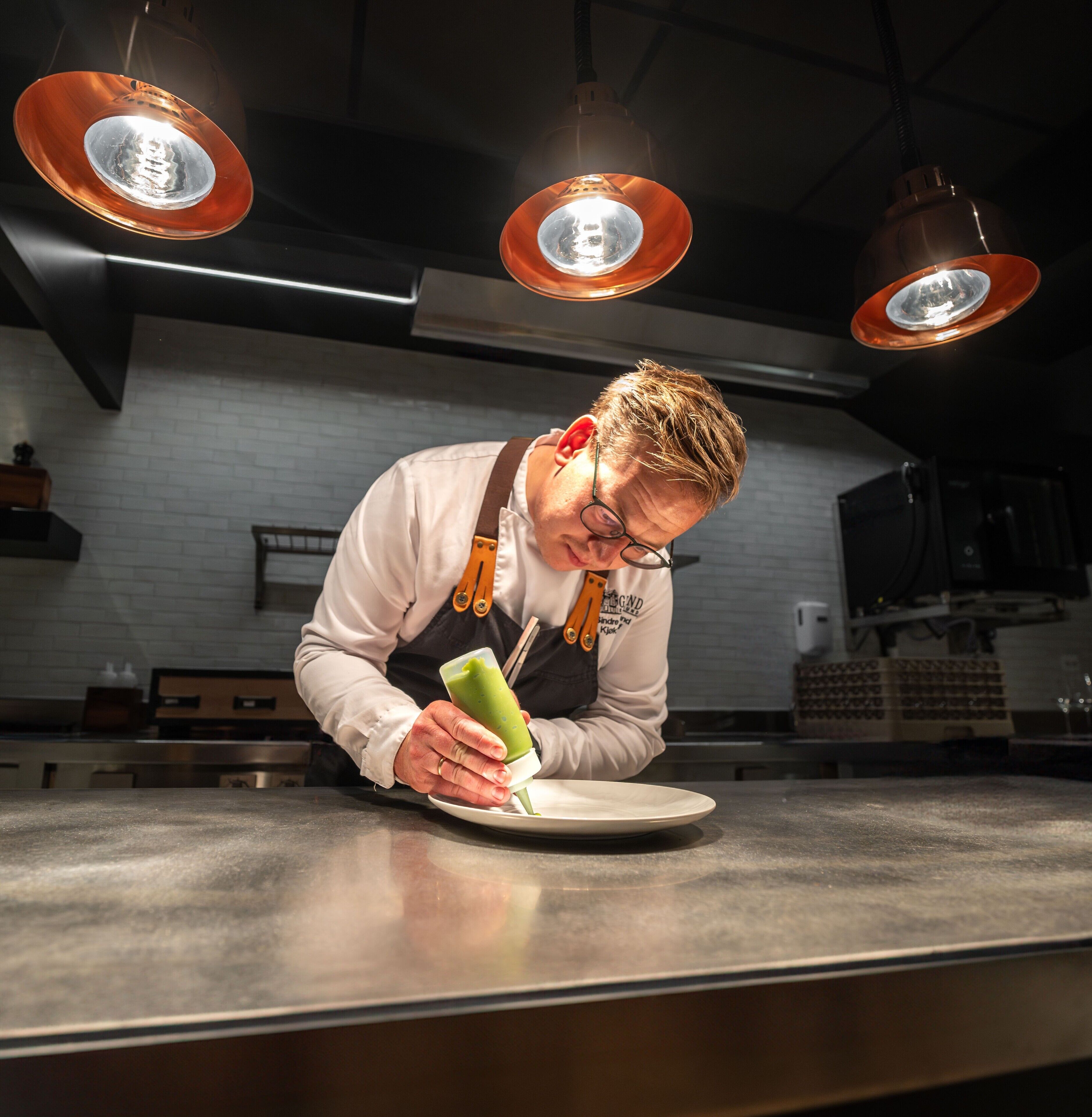Una persona preparando comida en una cocina con iluminación colgante y pared de ladrillo blanco.