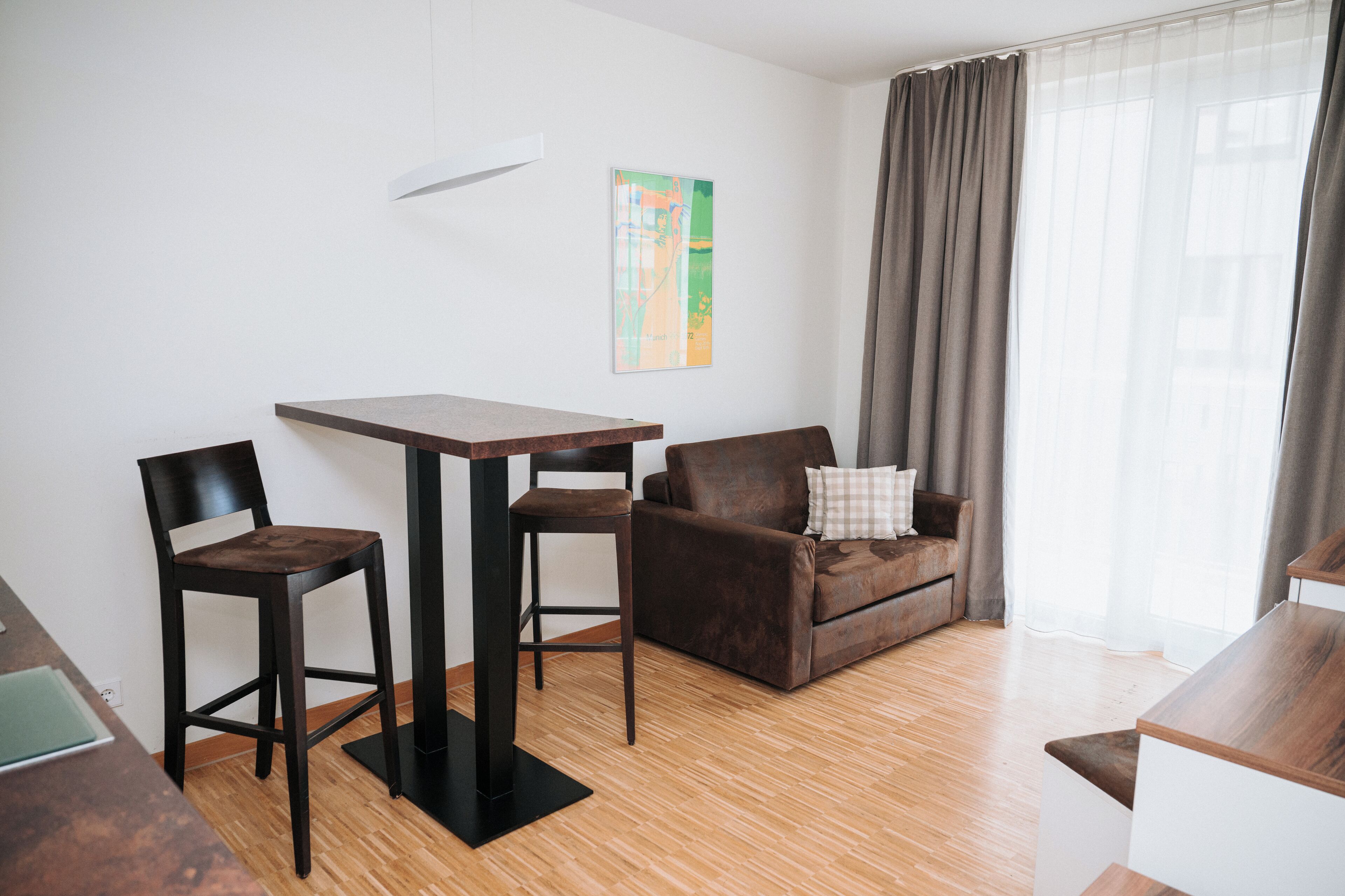 Una sala de estar moderna con una mesa de madera, dos sillas, un sofá marrón y un amplio ventanal con cortinas.