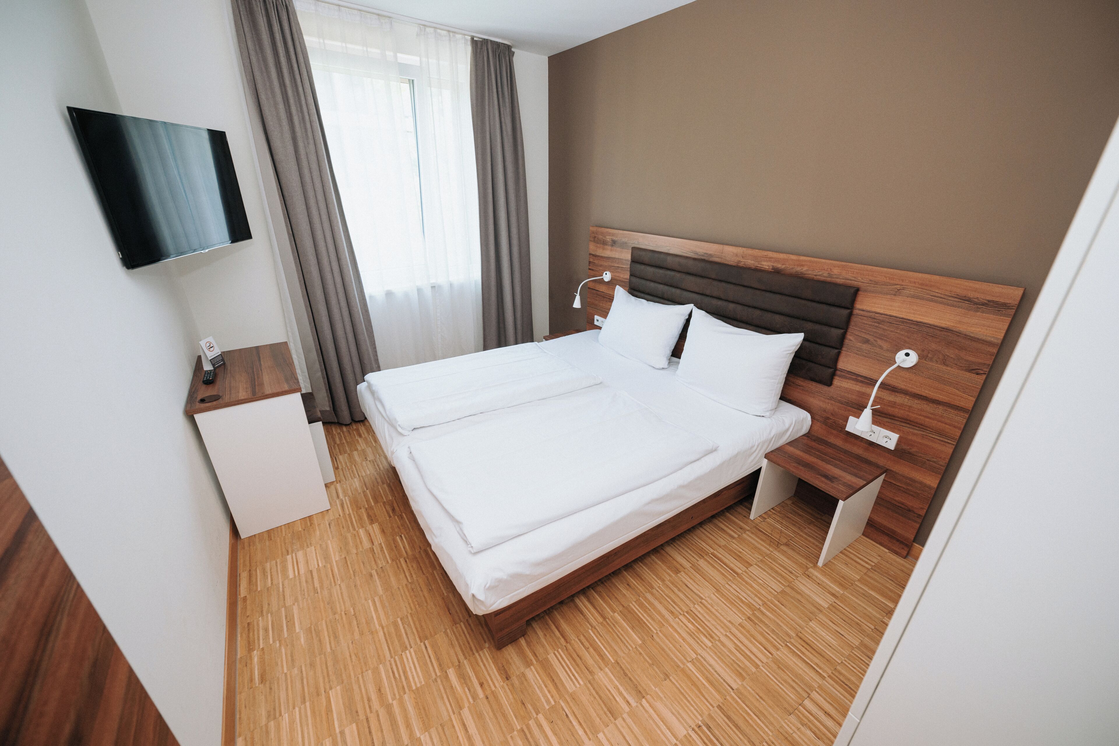 A hotel room with a large bed, a TV mounted on the wall, a bedside table, and a window with curtains.