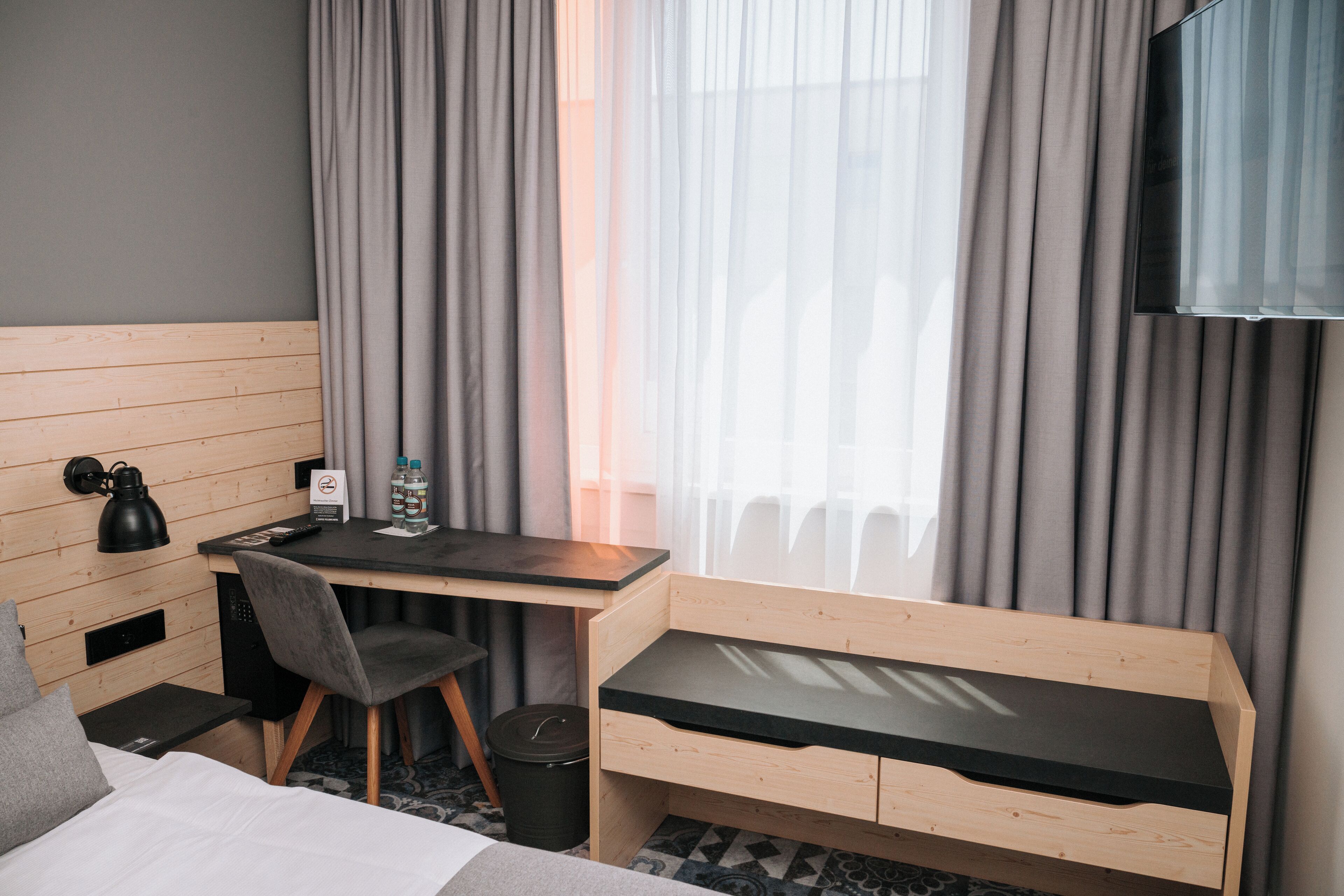 A hotel room with a bed, a desk with a chair, a TV, and a window with curtains.