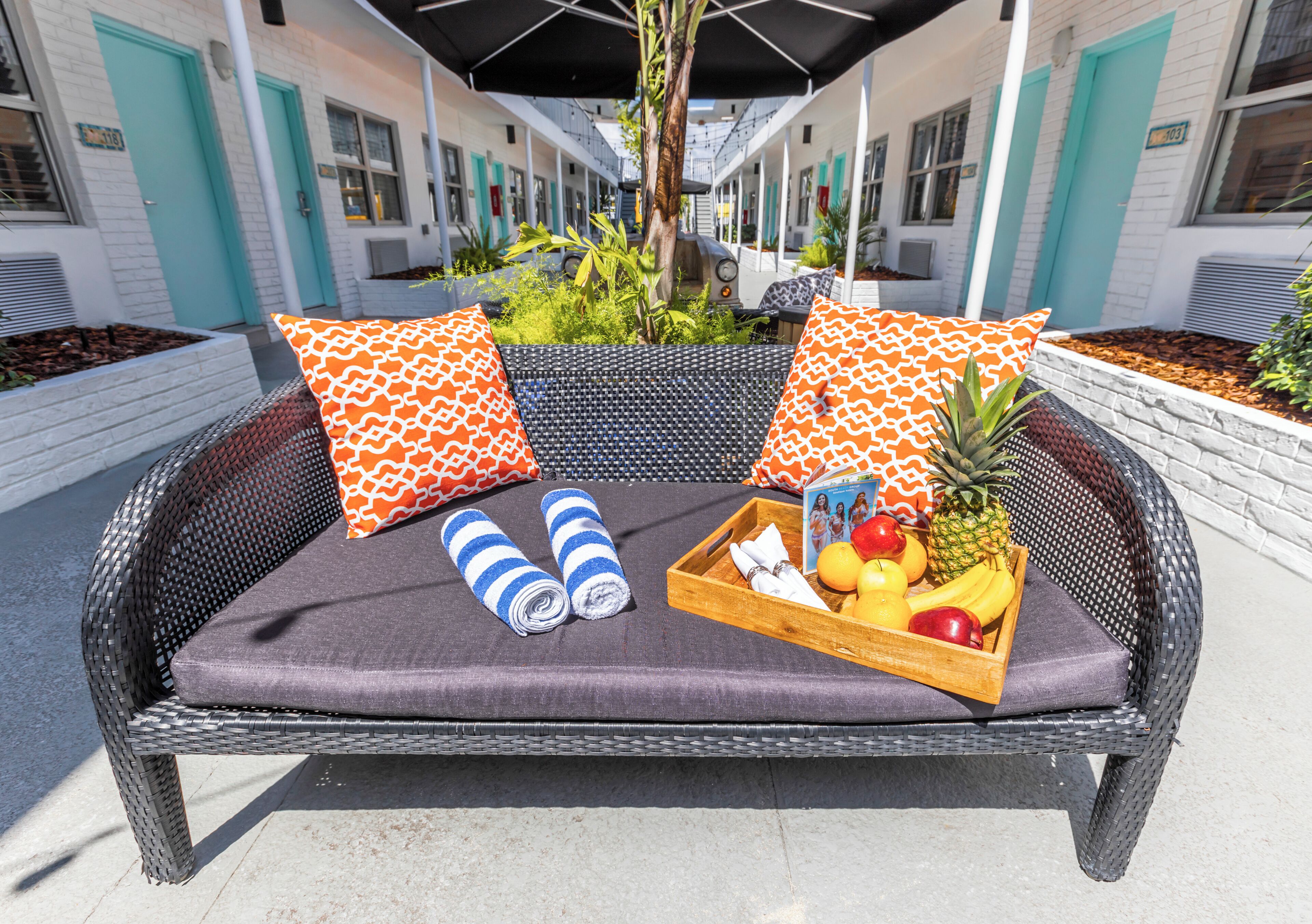 A wicker lounge chair with a cushion, two patterned pillows, a tray with fruits, and rolled towels.
