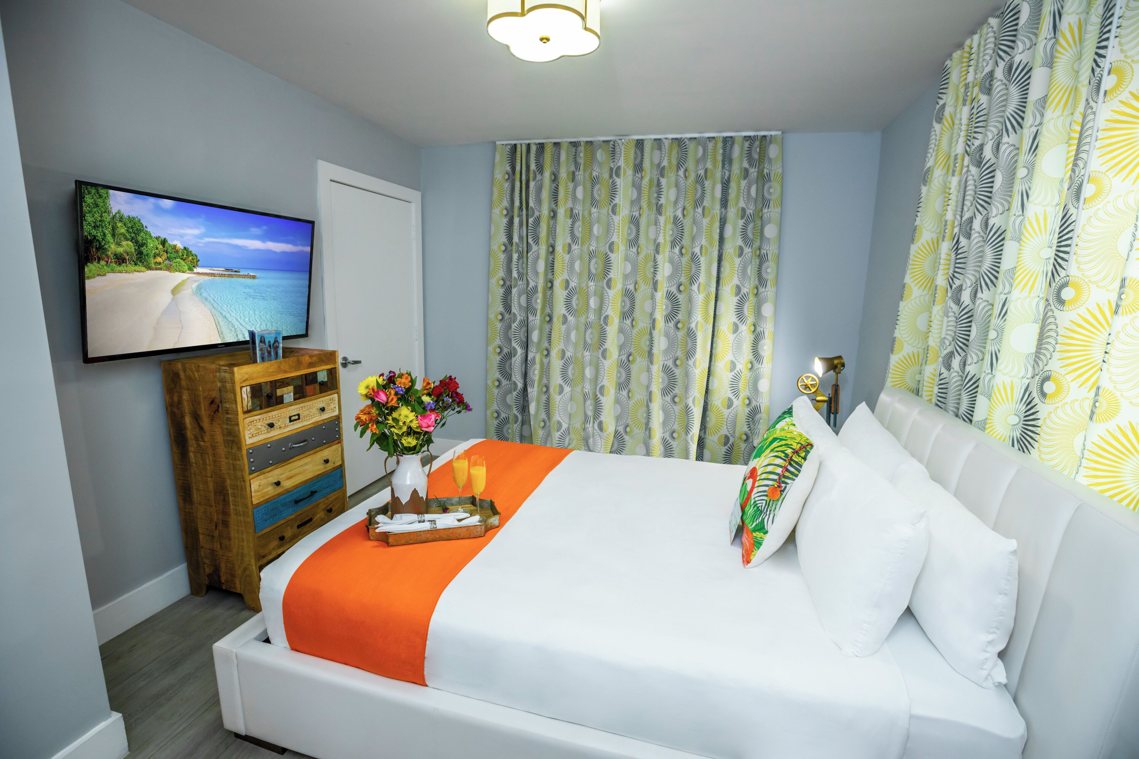 A hotel room with a bed, a TV mounted on the wall, a wooden dresser, and a window with patterned curtains.