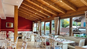 A dining area with wooden beams, white furniture, and large windows offering a view of the outside.