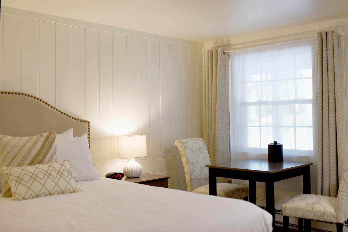 Habitación de hotel con una cama grande, dos sillas, una mesita, una lámpara y una ventana con cortinas.