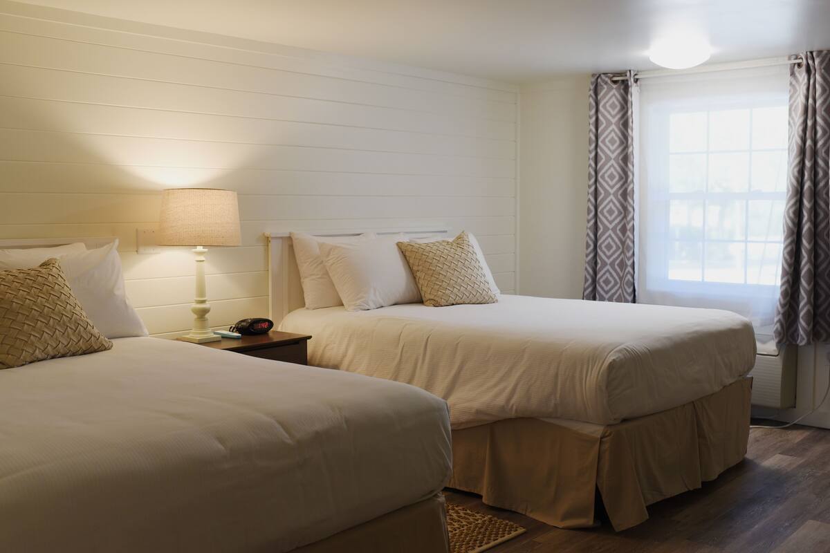 Una habitación con dos camas, una mesita de noche con lámpara y una ventana con cortinas.