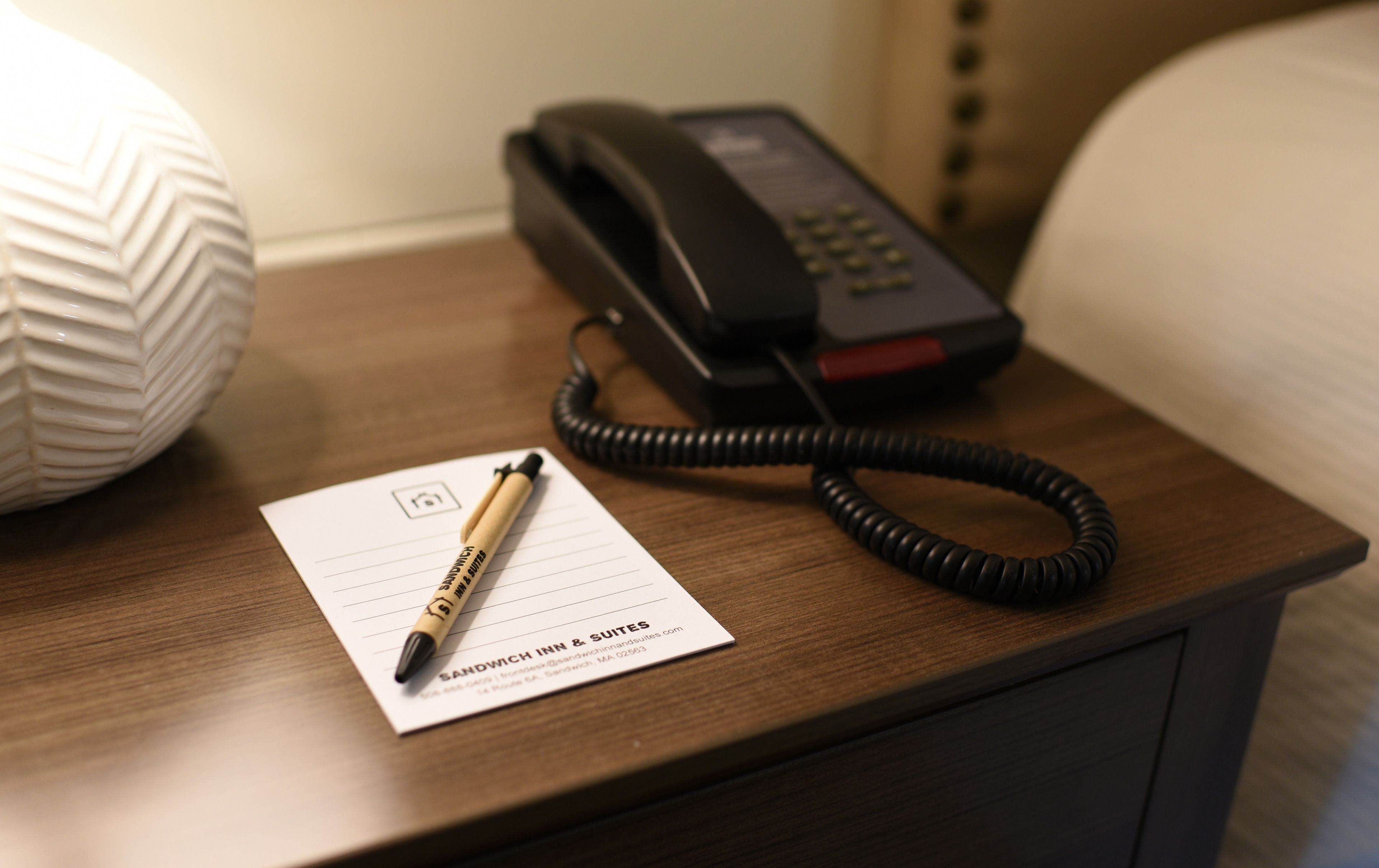 Een houten nachtkastje met een zwarte telefoon, een pen en een visitekaartje.