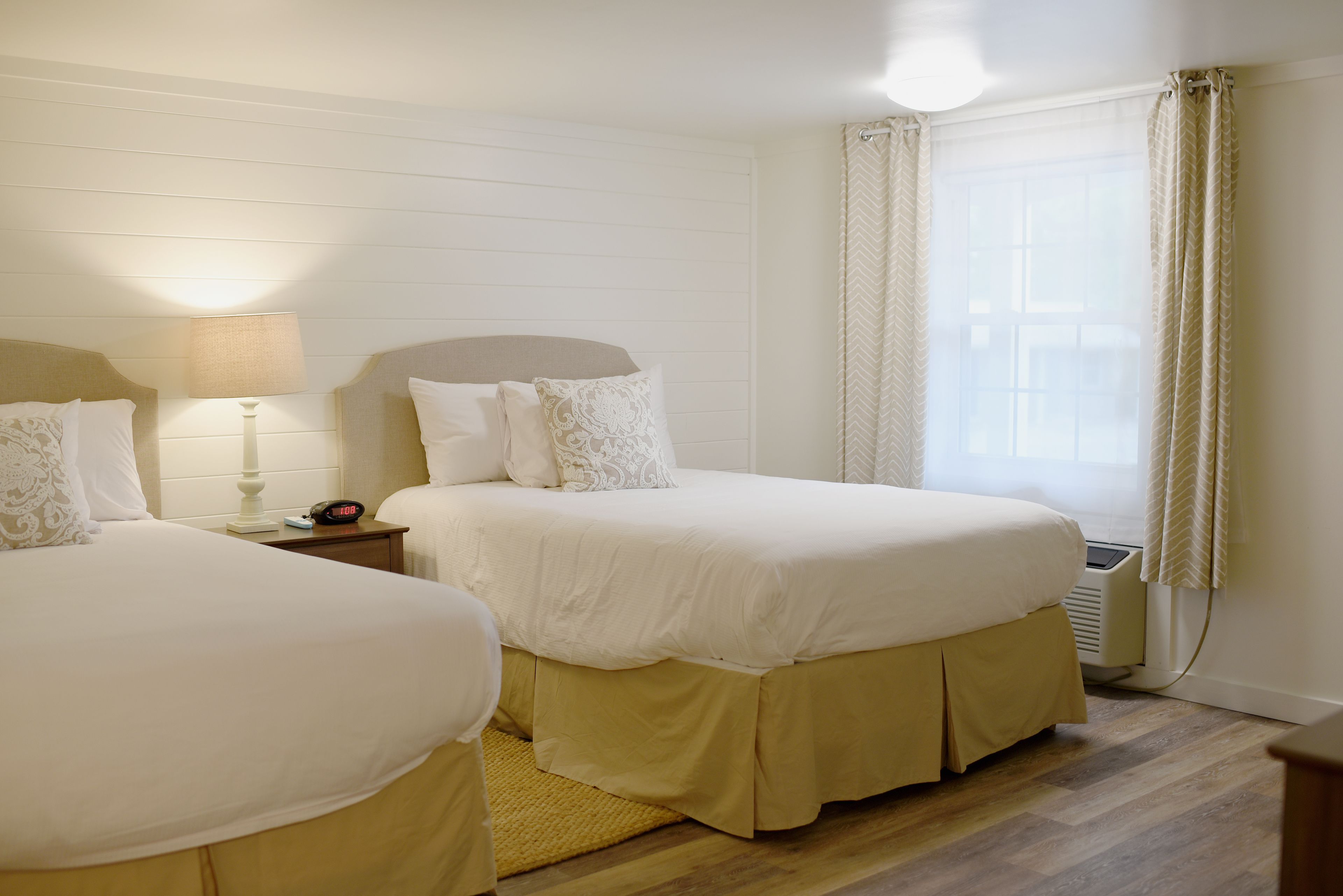 Une chambre d’hôtel avec deux lits, une table de chevet avec une lampe, une horloge et une fenêtre avec des rideaux.