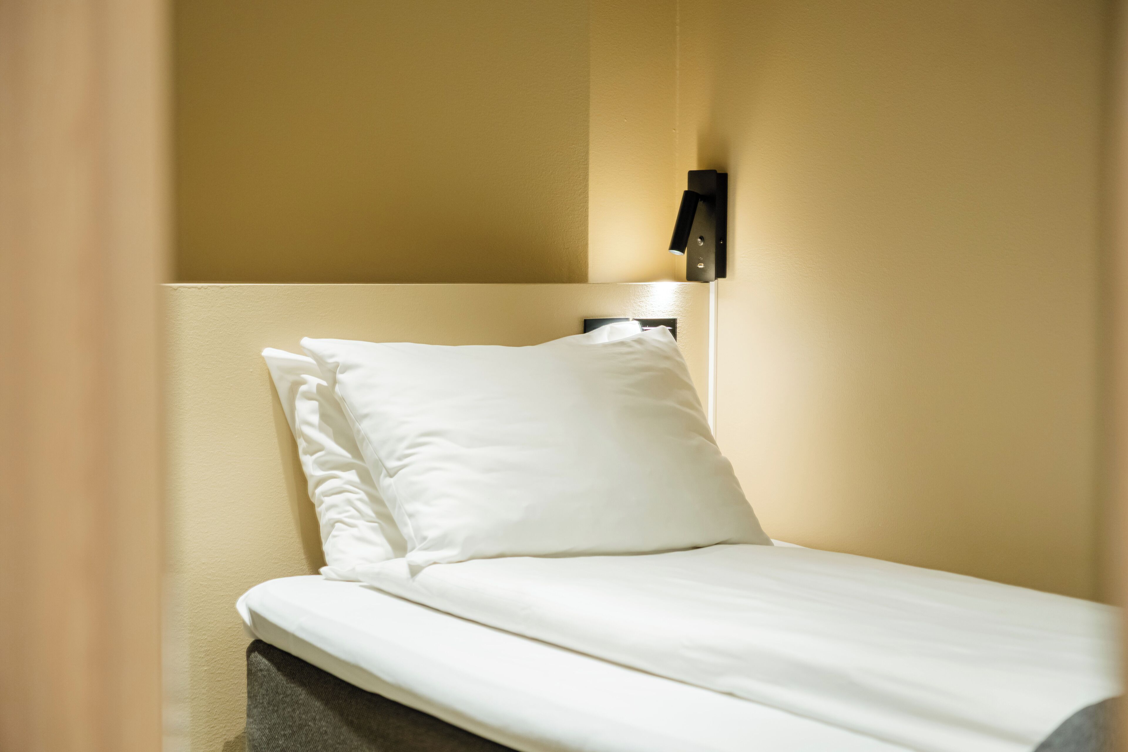 A neatly made bed with white bedding and a headboard, a wall-mounted light fixture, and a partial view of a beige wall.