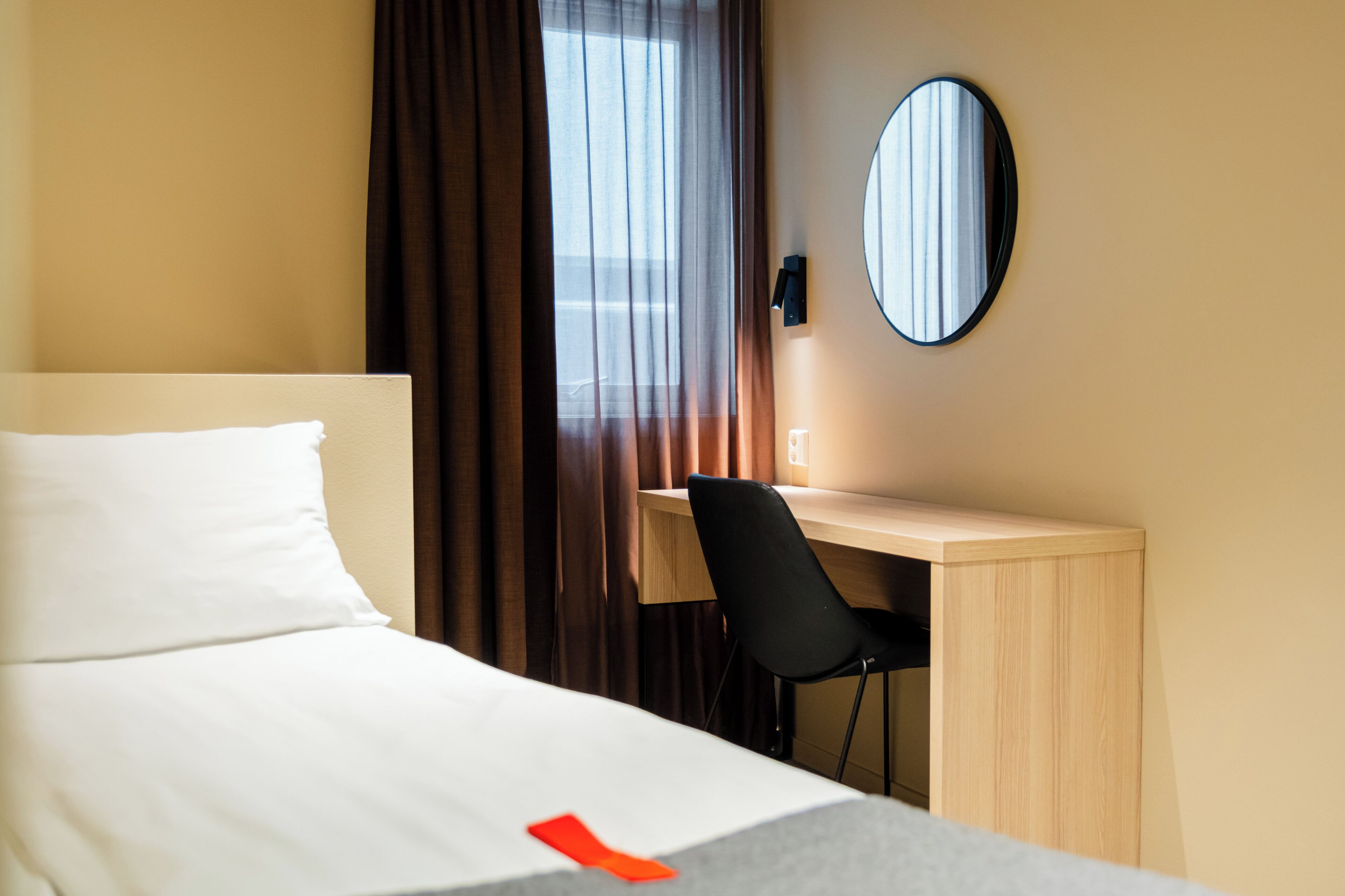 Une chambre d’hôtel avec un lit, un bureau, une chaise, un miroir rond et une fenêtre avec des rideaux.