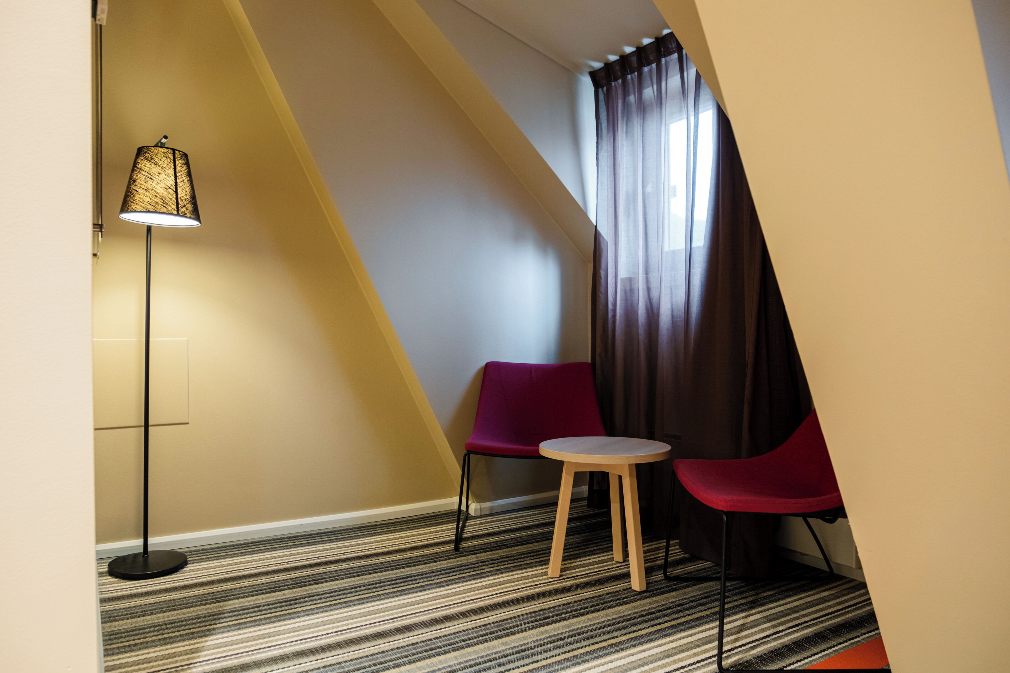 Habitación de esquina con alfombra a rayas, una lámpara de pie, dos sillones y una ventana con cortinas.