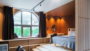 Et soveværelse med seng, skrivebord, stol, et vindue med gardiner og udsigt ud.