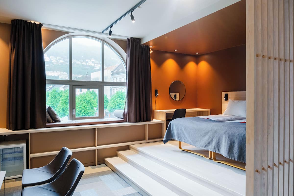Un dormitorio con cama, escritorio, silla, ventana con cortinas y vistas al exterior.