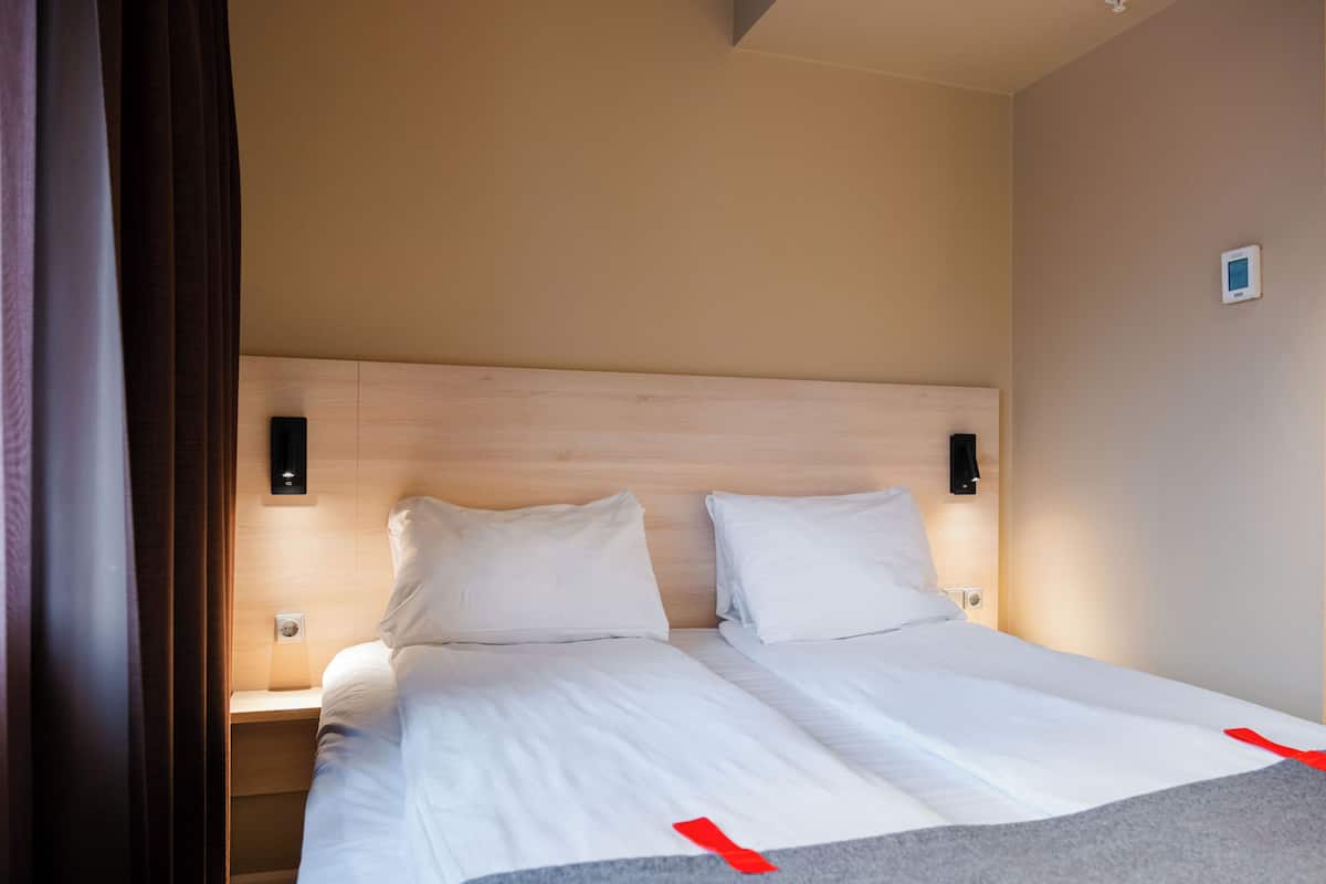Habitación de hotel con cabecera de madera, dos camas blancas, una mesita de noche y un termostato instalado en la pared.