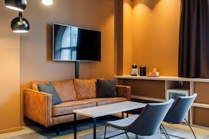 Ein modernes Wohnzimmer mit einer Couch, einem Couchtisch, zwei Sesseln, einem flachbildfernseher an der Wand und einem kleinen Regal mit Dekorationsobjekten.