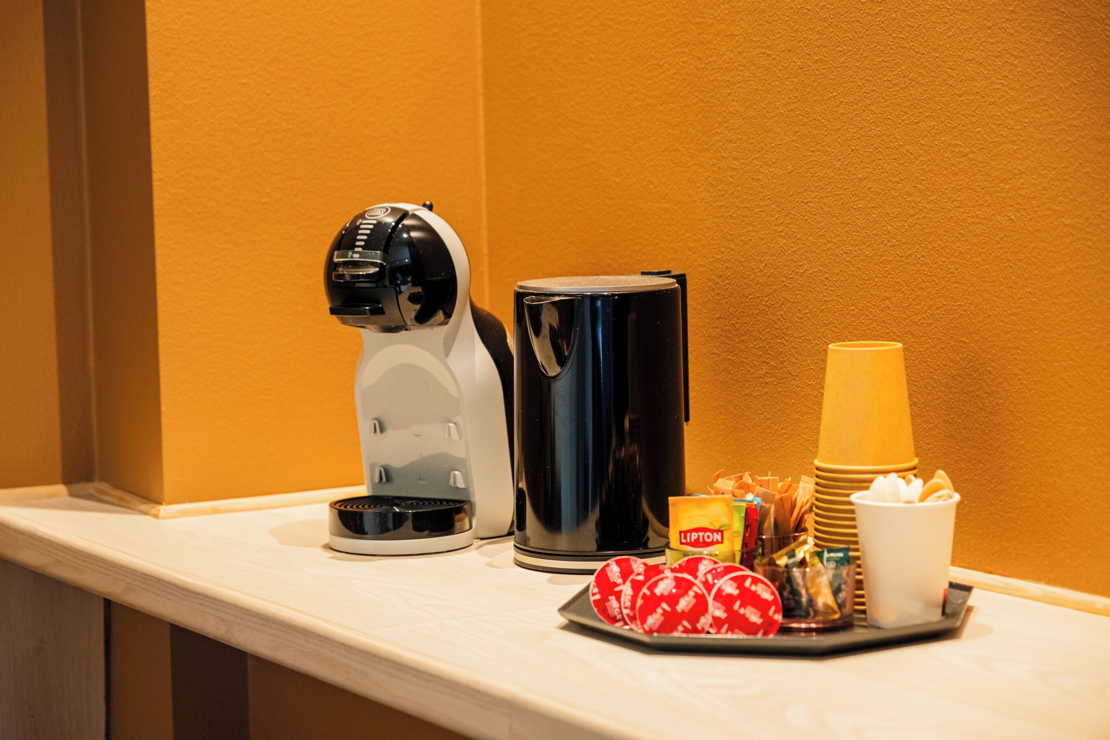 Uma cafeteira, um bule, copos descartáveis e uma bandeja com petiscos em uma bancada de hotel.