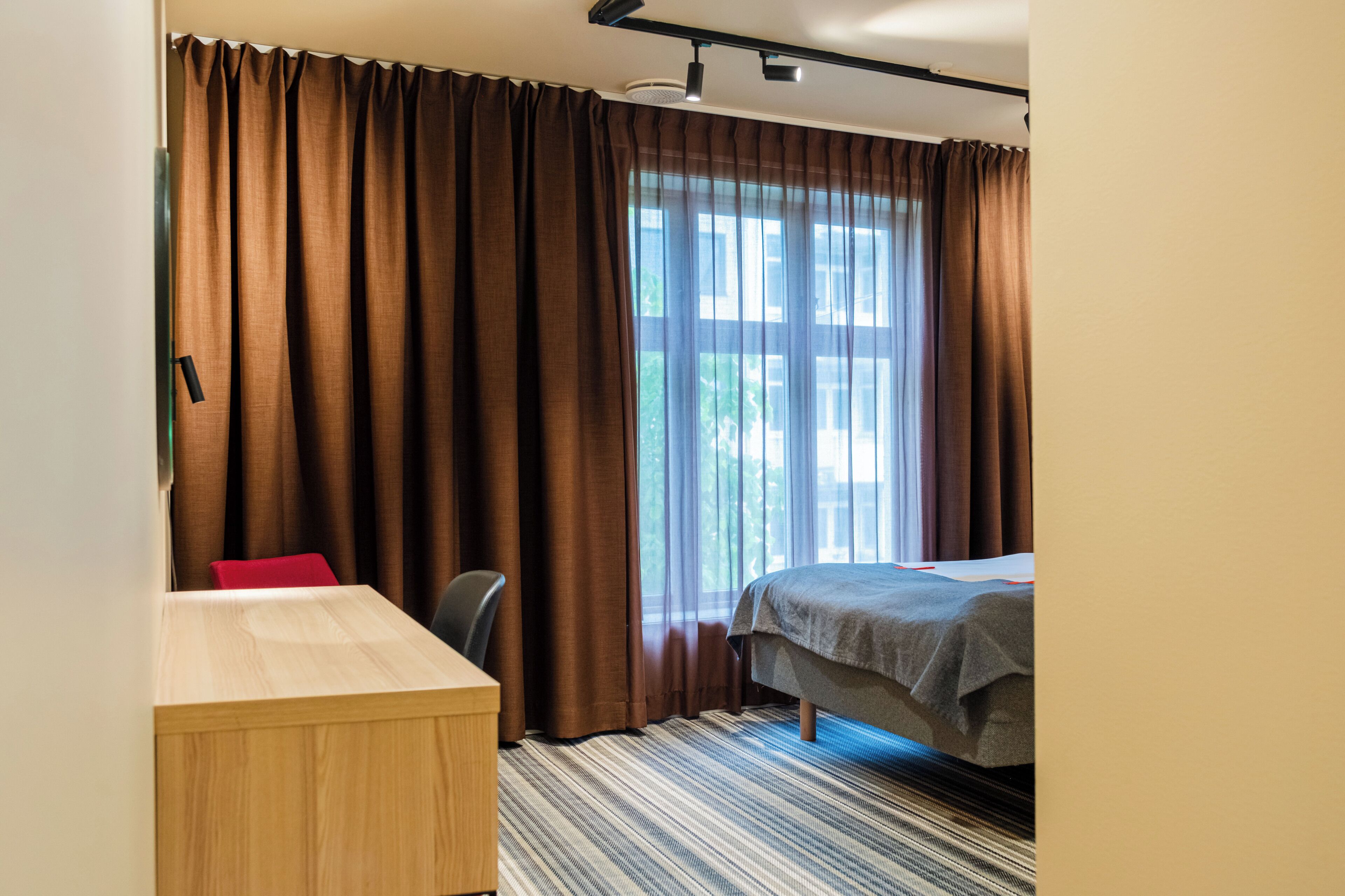 A hotel room with a bed, a desk, a chair, and a large window with curtains.