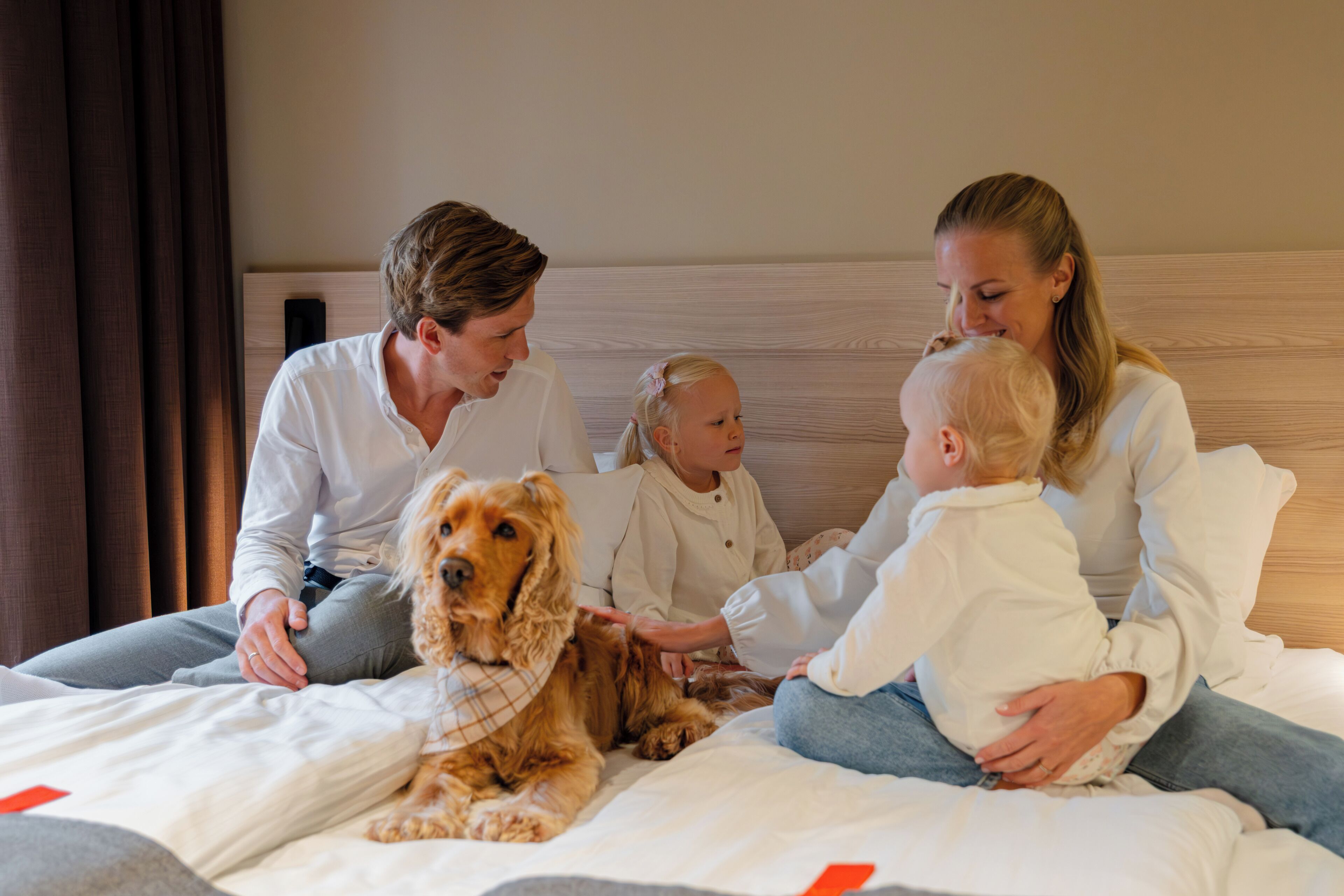 Una familia con un perro en una cama dentro de una habitación de hotel.