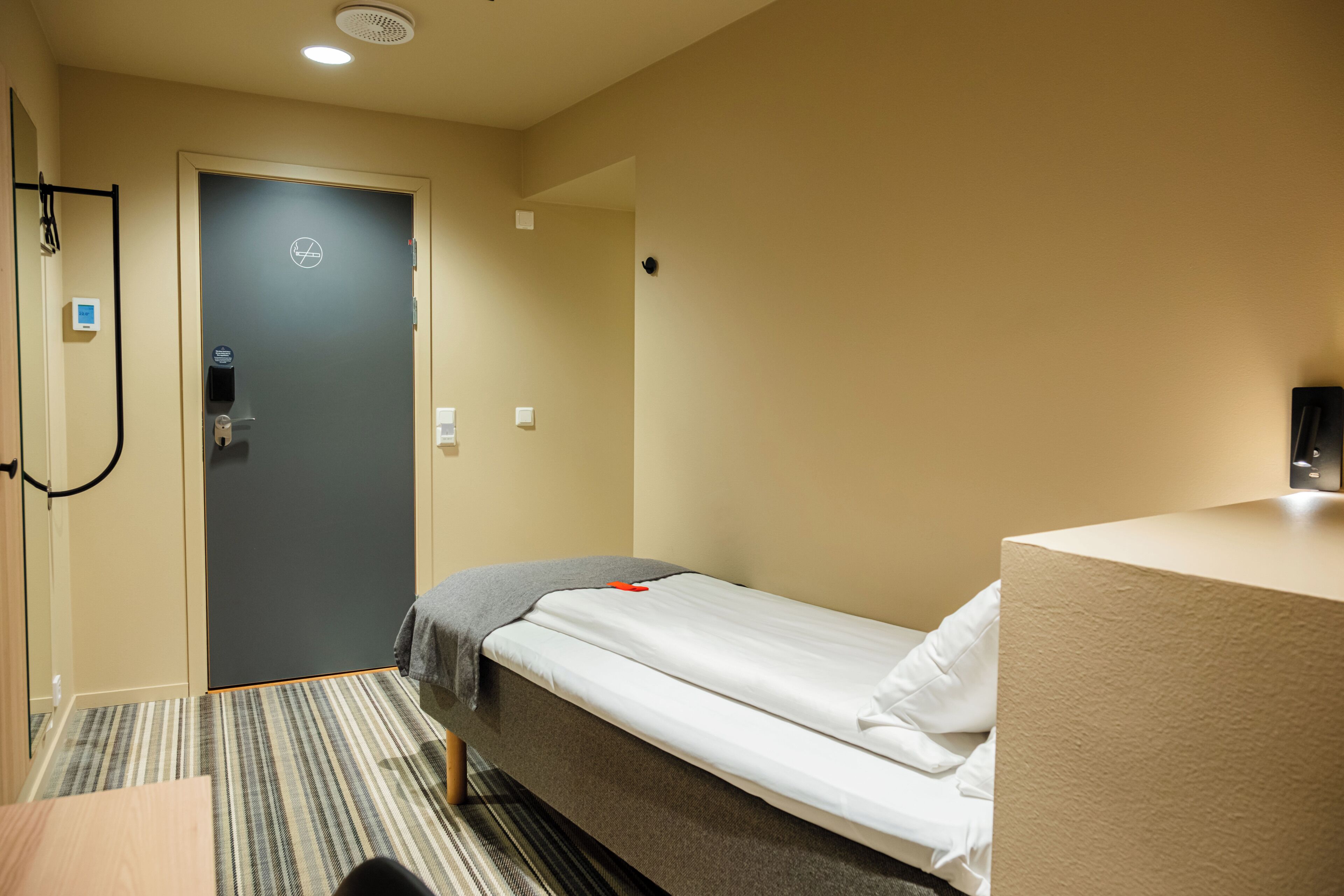 A hotel room with a bed, a desk, a chair, a door, and a wall-mounted light.