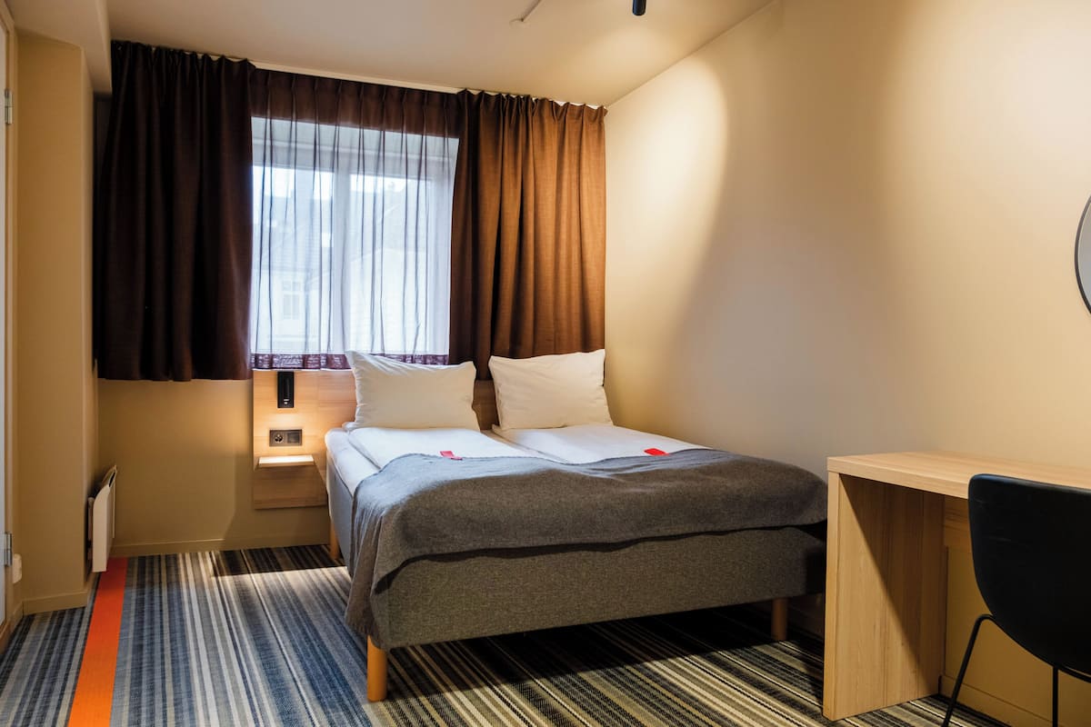 Habitación de hotel con cama, escritorio, silla, ventana con cortinas y radiador.