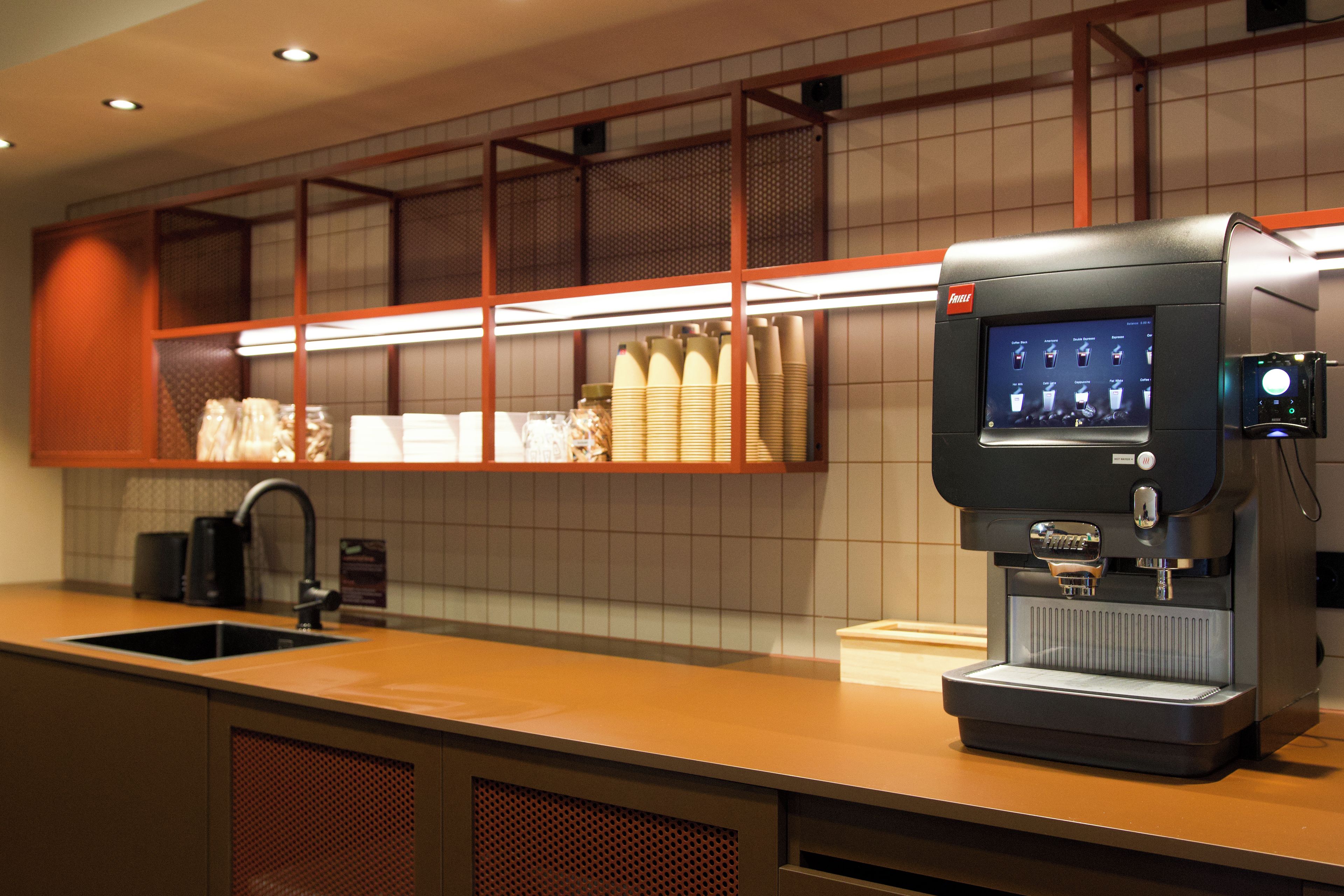Una cafetera con pantalla digital, un lavamanos y un salpicadero de azulejos.