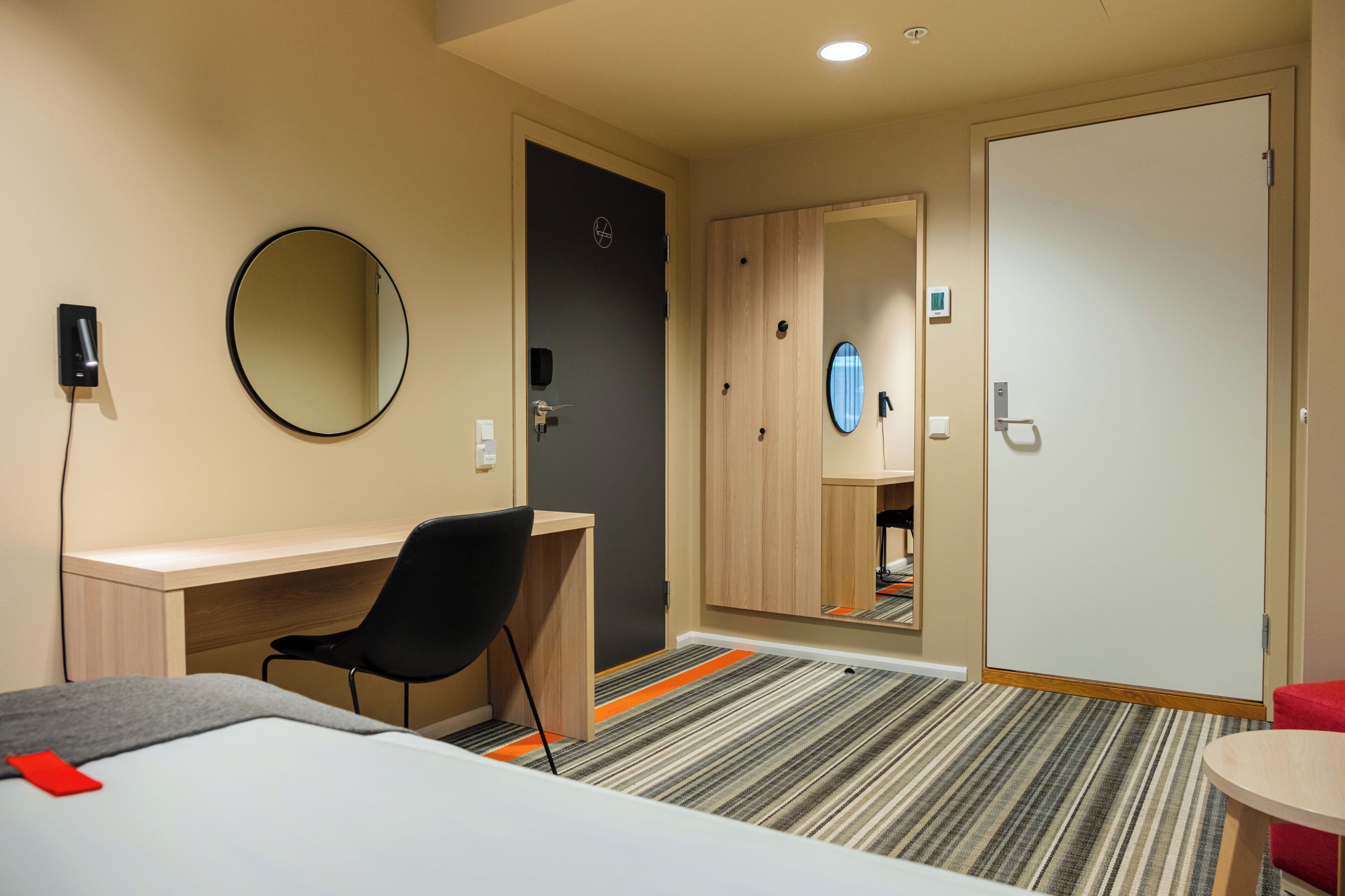 A hotel room with a bed, a desk, a chair, a round mirror, a wardrobe, and a small table.