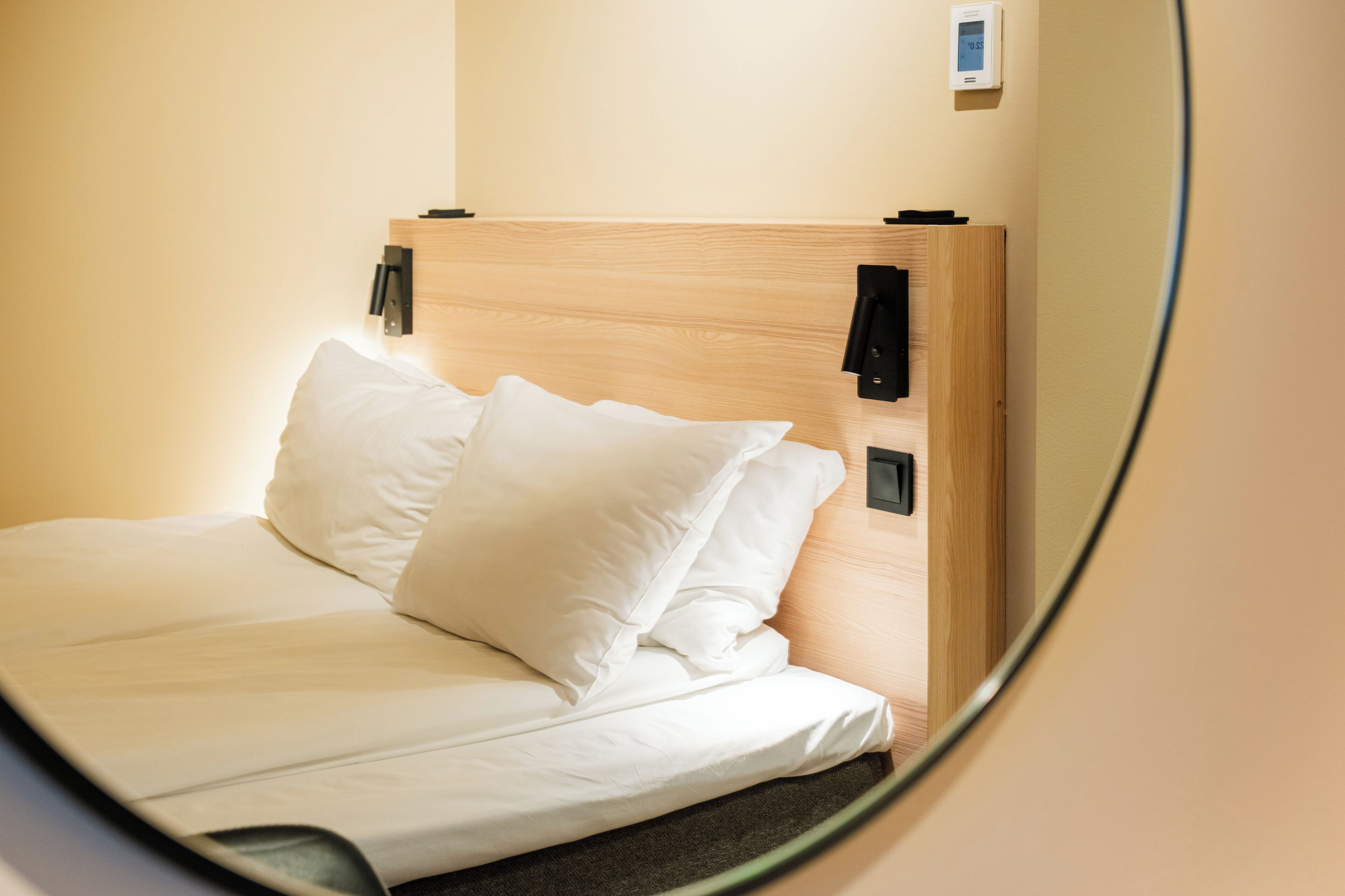 A neatly made bed with white linens and pillows, a wooden headboard, and a wall-mounted thermostat.