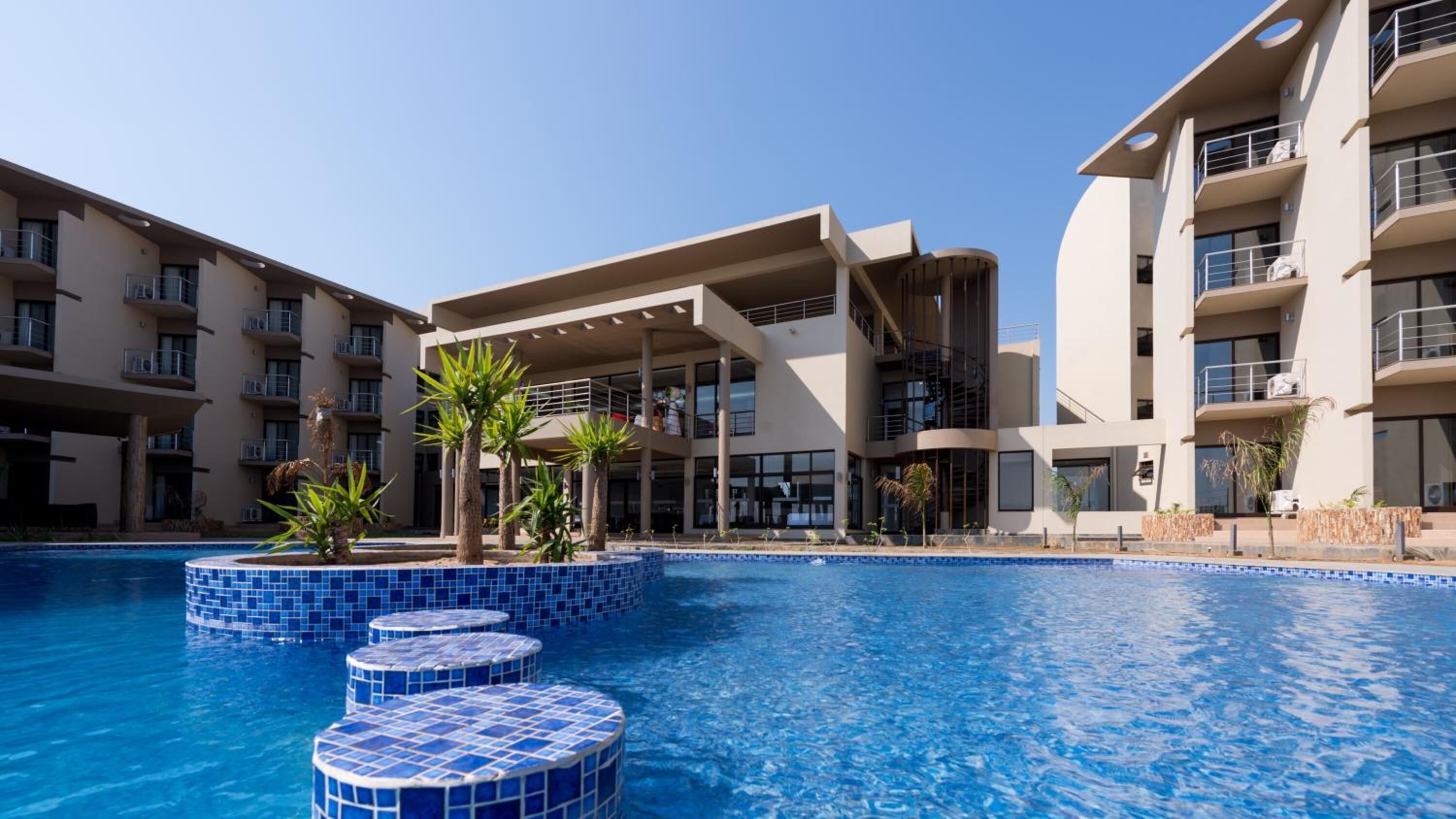 A modern hotel with a swimming pool, multiple balconies, and a central open-air area with palm trees.