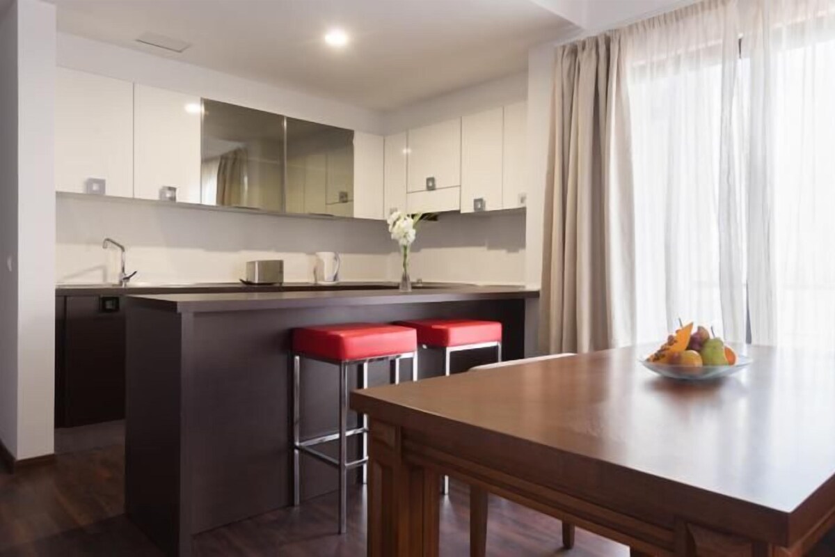 Una cocina moderna con una isla central, taburetes de bar, una mesa de comedor y un frutero.
