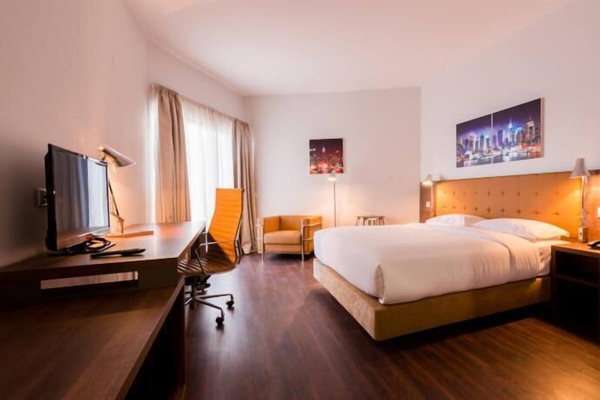 A hotel room with a bed, a desk with a computer, a chair, a TV, and a nightstand.