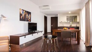 A modern living room with a flat-screen TV, a dining area with a wooden table and bar stools, and a kitchen with a sink and countertop.