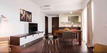 Una sala de estar moderna con un televisor de pantalla plana, una zona de comedor con mesa de madera y taburetes altos, y una cocina con pileta y encimera.